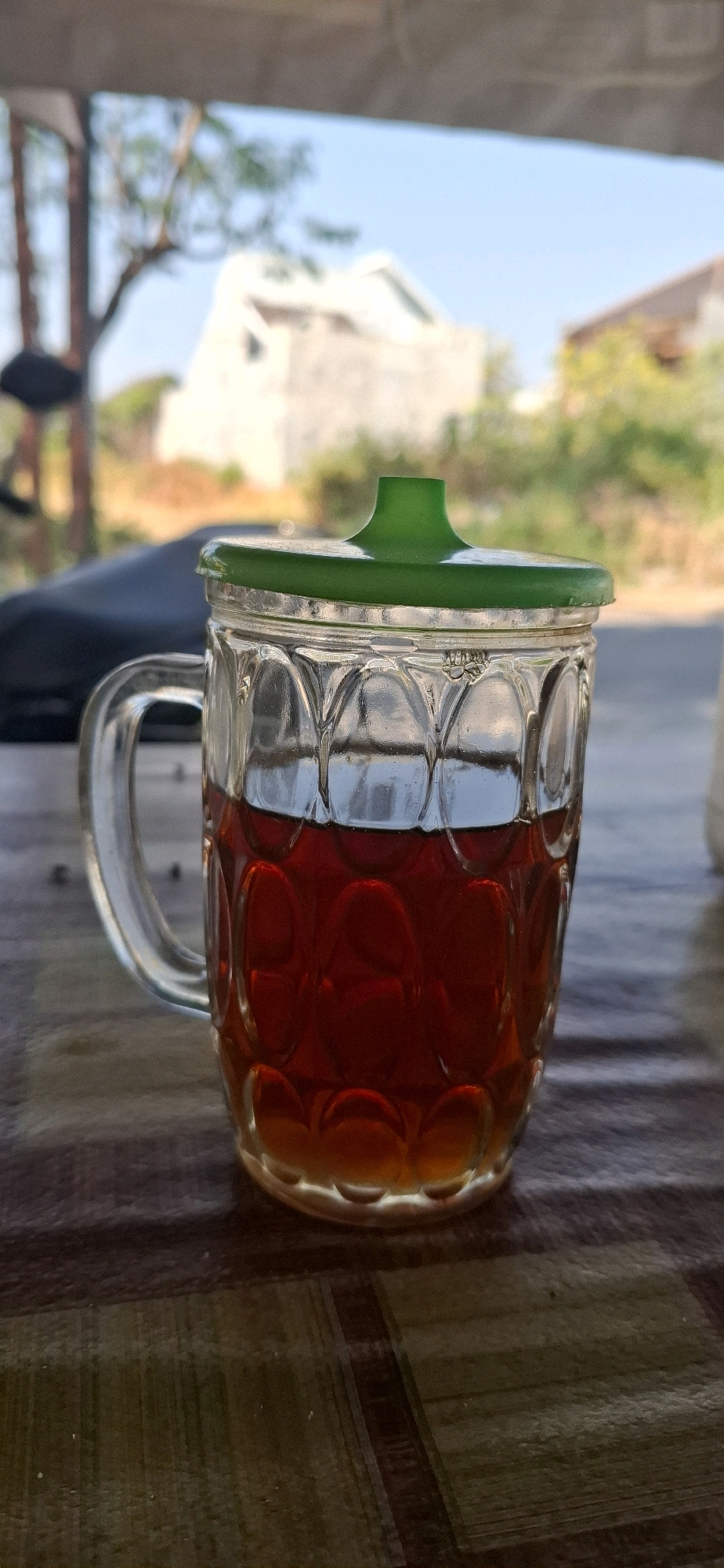 A glass mug filled with a dark amber liquid, likely tea, sits on a patterned surface. The mug has a clear glass body with a textured design and a clear handle. A bright green lid with a raised knob covers the top of the mug. In the background, out of focus, are buildings, trees, and a clear blue sky, suggesting an outdoor setting in a city like Gedongan, Indonesia. The lighting indicates it is daytime.