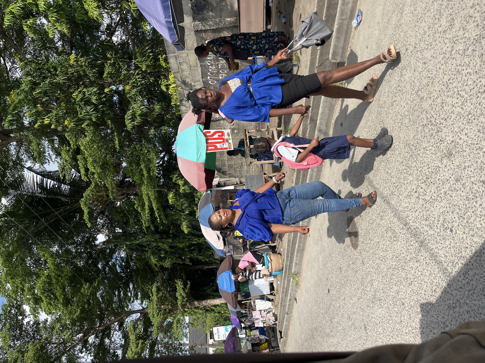Two women and a child are walking on a concrete surface. One woman wears a blue top and jeans, and the other wears a blue top and black skirt. The child wears a blue top and shorts, and a pink backpack. Trees are visible in the background on the left. Several umbrellas are visible with people under them. There is a POS sign visible next to one of the umbrellas. This scene is in Ado, Nigeria.