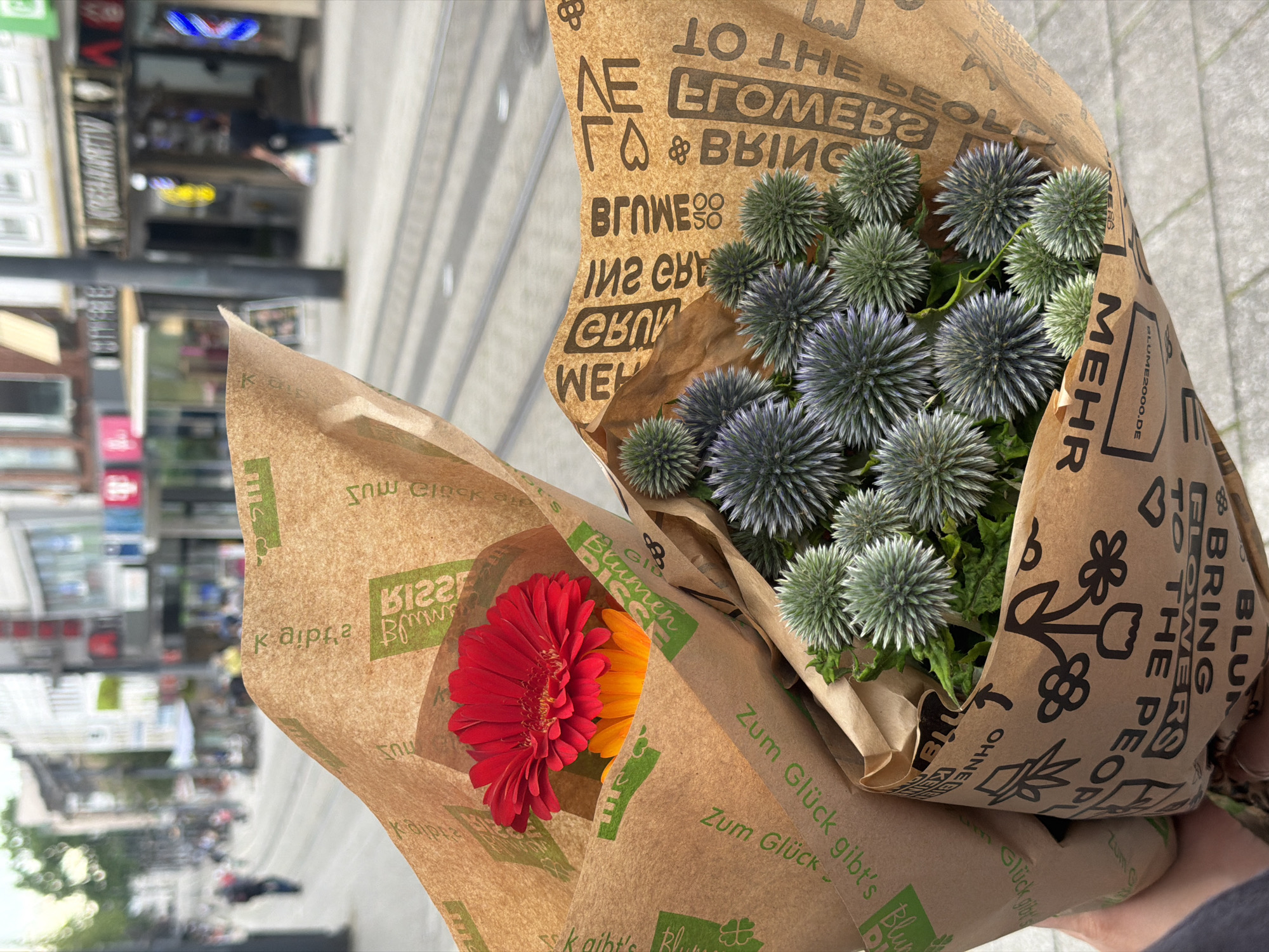 The image displays two bouquets of flowers wrapped in brown paper, held by a person's hands in the foreground. The bouquet on the left contains a bright red Gerbera daisy and an orange flower beneath it. The bouquet on the right features numerous spherical, blue-green flowers with spiky petals. Both bouquets are wrapped in brown paper with green lettering and decorative motifs. In the background, a blurred city street scene is visible, with buildings, shops, and a few pedestrians. The presence of tram tracks on the street indicates an urban environment. The shops have illuminated signs. One sign reads "KREMHANATIV". The overall atmosphere suggests a casual, outdoor setting in a German city. The style of the shops and the German text on the wrapping paper suggest the location is likely Witten, Germany.