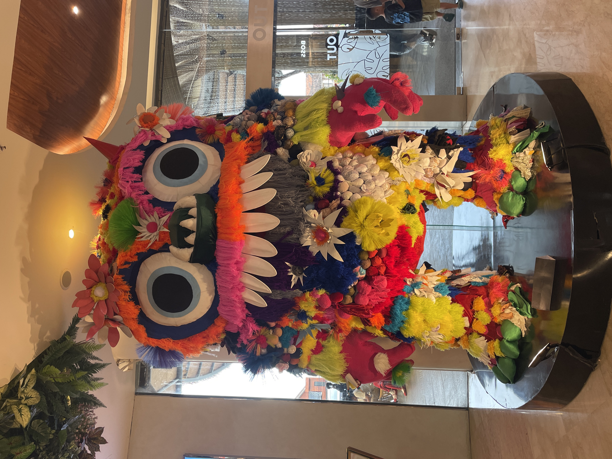 The image depicts a large, colorful, and fantastical sculpture resembling a creature or monster, possibly a display or art installation. The sculpture is highly ornamented with an array of textures, including yarn, flowers, fabrics, and other decorative materials. It features oversized eyes, large teeth, and various protrusions, suggesting a whimsical and somewhat menacing character.
The sculpture is standing on a polished black, circular base, which is placed on a light-colored tile floor. Behind the sculpture is a glass facade, reflecting the scene and also revealing an exterior view of people standing or walking outside. The glass door has "OUT" and "TUO" written on it.
Based on the architectural style and branding elements, the sculpture is located inside a commercial building, potentially a mall or retail space, with modern interior design elements.
The image is taken at the 'Tu Outlet' store, located in Denpasar, Indonesia.