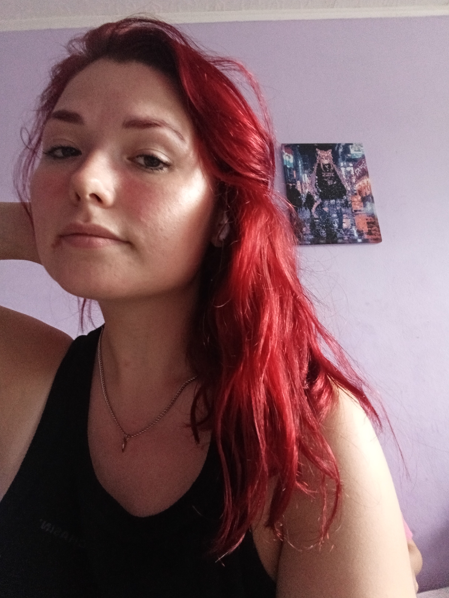 A young woman with bright red hair is shown from the chest up, looking at the camera. She is wearing a black tank top and a silver chain with a small pendant. Her left arm is bent and her hand rests on her chin. The background is a plain purple wall with a small framed picture of a cat on it. The lighting suggests the photo was taken indoors, possibly during the day, with natural light coming from the left.