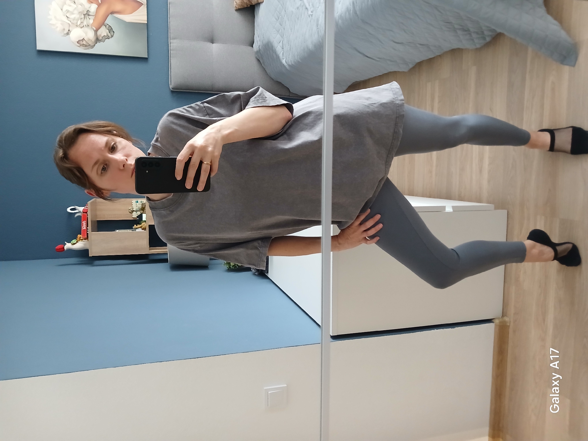 A person is seen in a mirror selfie in an interior setting. The person is wearing a grey oversized t-shirt and grey leggings, along with black footwear. The environment includes a blue wall, a white cabinet or wardrobe, and a shelf with decorations. There is also a bed visible in the reflection. The location is suggested to be Yekaterinburg, Russia. However, there is no evidence in the image that supports the location.