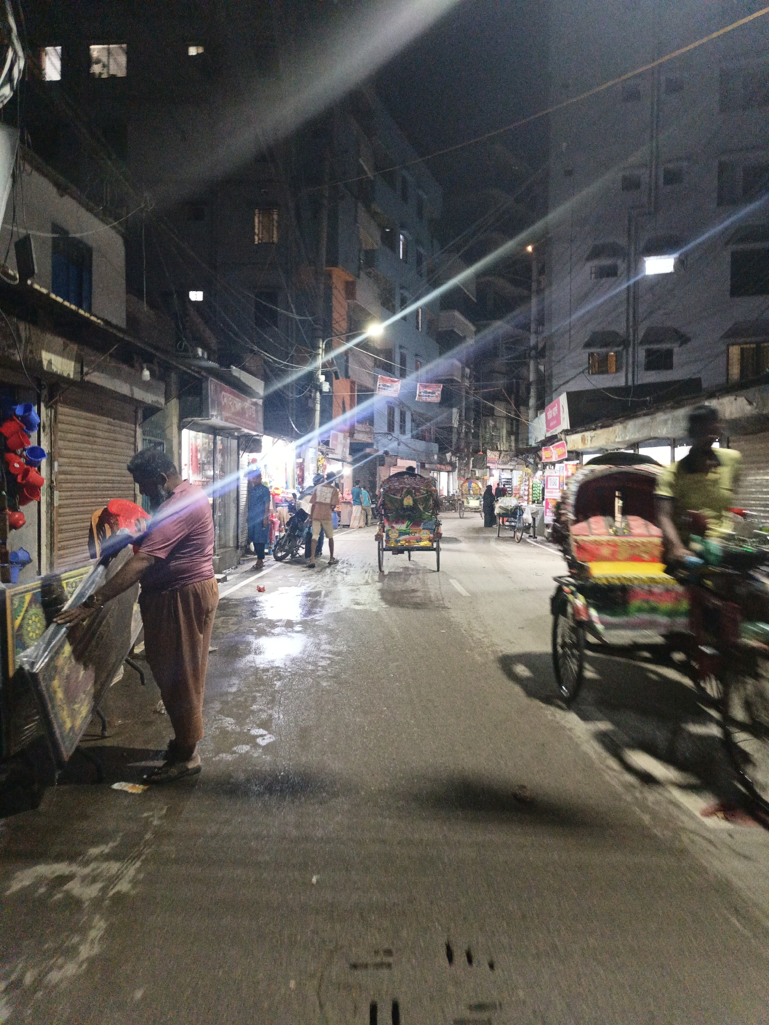The image depicts a street scene at night, likely in a bustling urban area. The street is wet, reflecting the ambient light. On the left, a man in a mauve shirt and brown pants is seen attending to an object placed on a stand. Several people are visible further down the street, along with parked motorcycles and bicycles. In the foreground, a rickshaw is blurred, indicating movement. Other rickshaws are visible down the street. The buildings lining the street are multi-storied and have various shops and residences. Above, a network of utility wires is strung between the buildings. The scene is illuminated by streetlights, which create streaks in the image due to the camera’s exposure.
