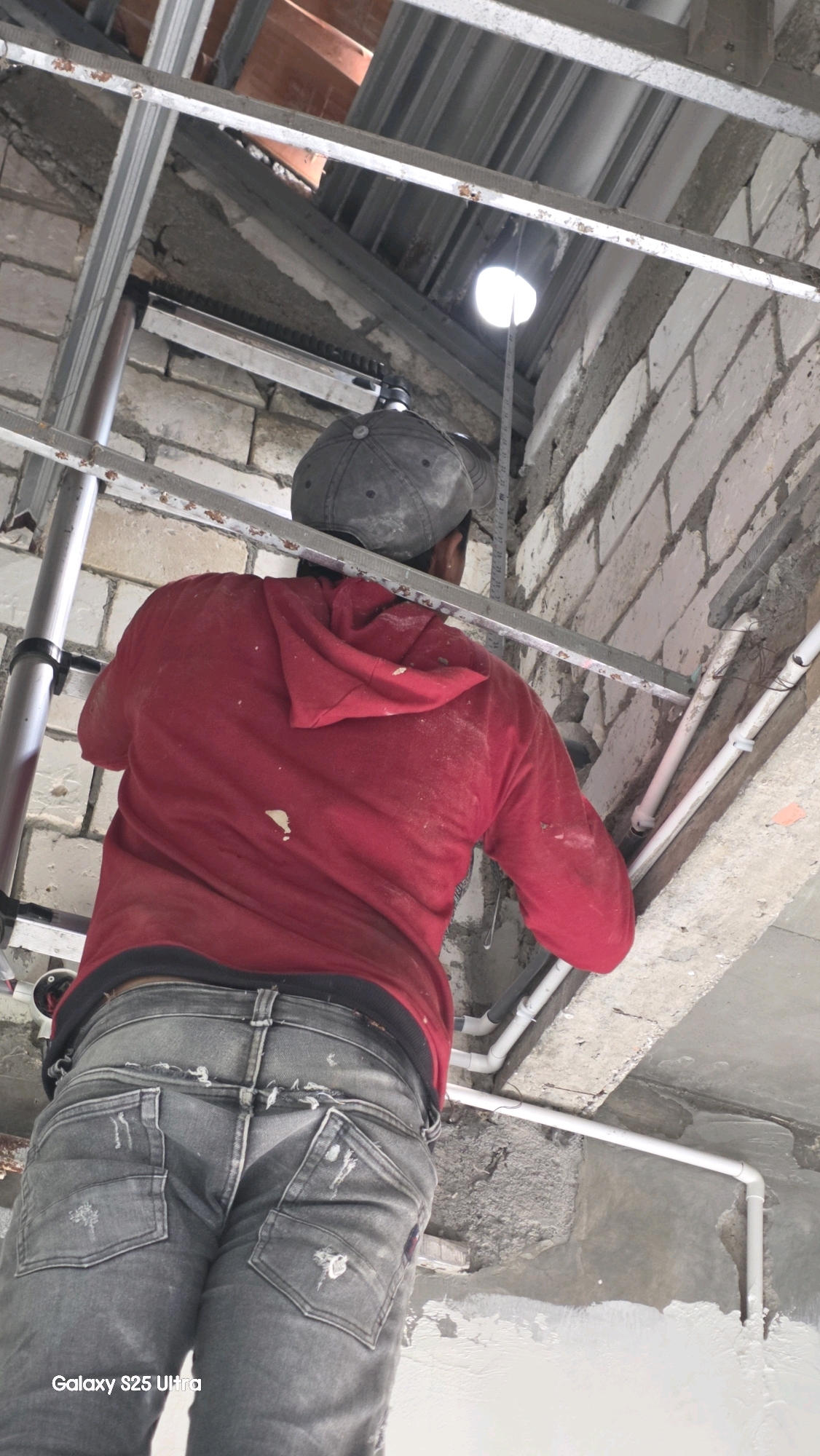 A construction worker, wearing a red hooded sweatshirt and grey distressed jeans, is working on a ceiling installation in Juwana, Indonesia. The worker, seen from behind, is holding a tape measure and appears to be measuring a distance from a light source on the ceiling structure. The environment is a construction site, with exposed brick walls, metal framing for the ceiling, and white pipes running along the ceiling. Dust is visible on the worker's clothing and the surrounding surfaces. The overall scene depicts active construction work in progress.
