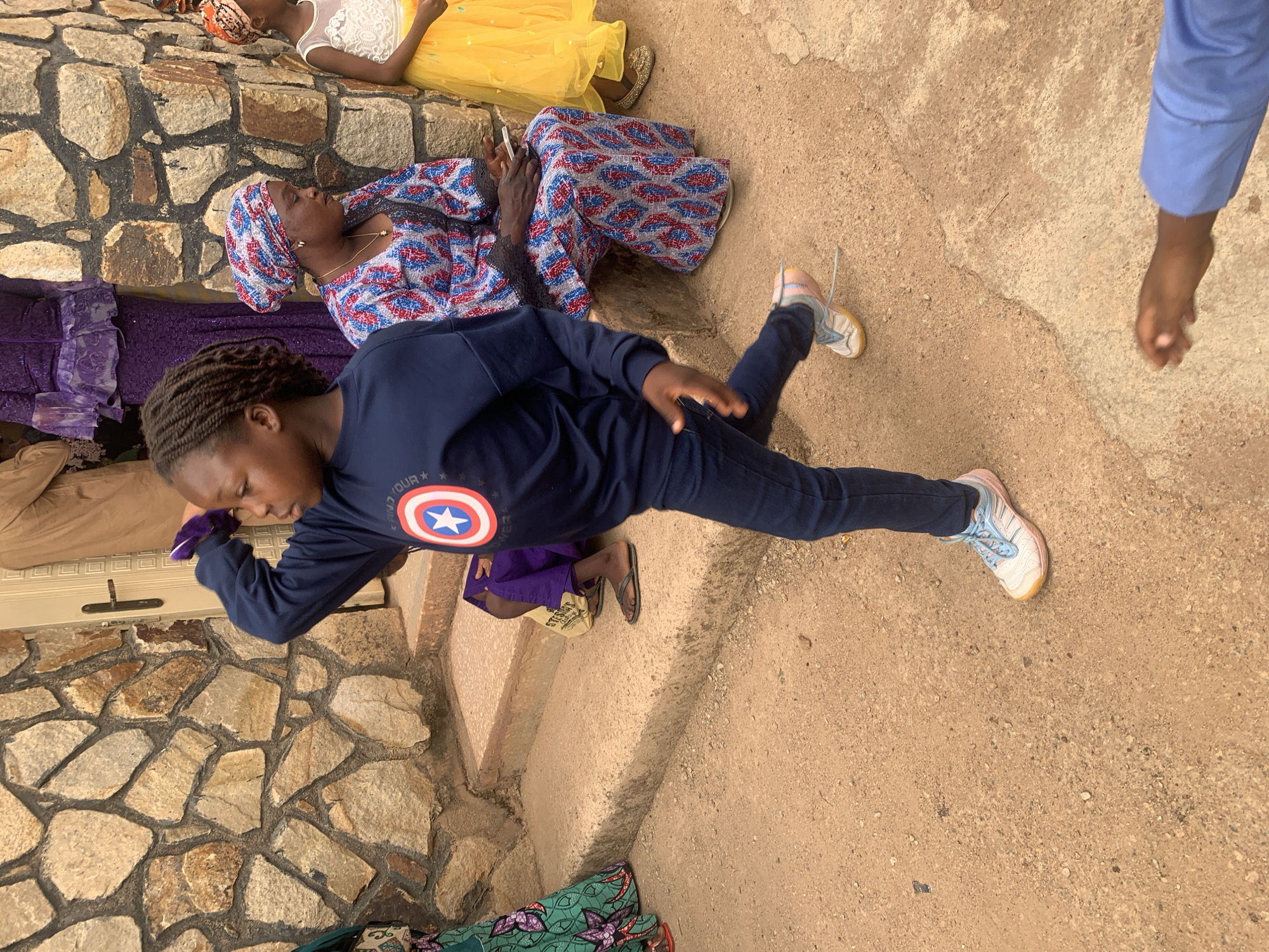 The image shows a group of people in what appears to be an outdoor gathering or resting area in Jos, Nigeria, based on the appearance of the environment. A young person wearing a dark blue long-sleeved shirt with a Captain America logo and blue jeans is lying on the ground, positioned with their head near stone steps and their feet extended outwards. Several other individuals are also present. One woman, wearing a blue and red patterned headscarf and clothing, is lying down, while another is dressed in yellow and white. Other people are partially visible. The ground is a light brown, and a stone wall or facade is present. The overall impression is one of relaxation or waiting in a public or semi-public space.