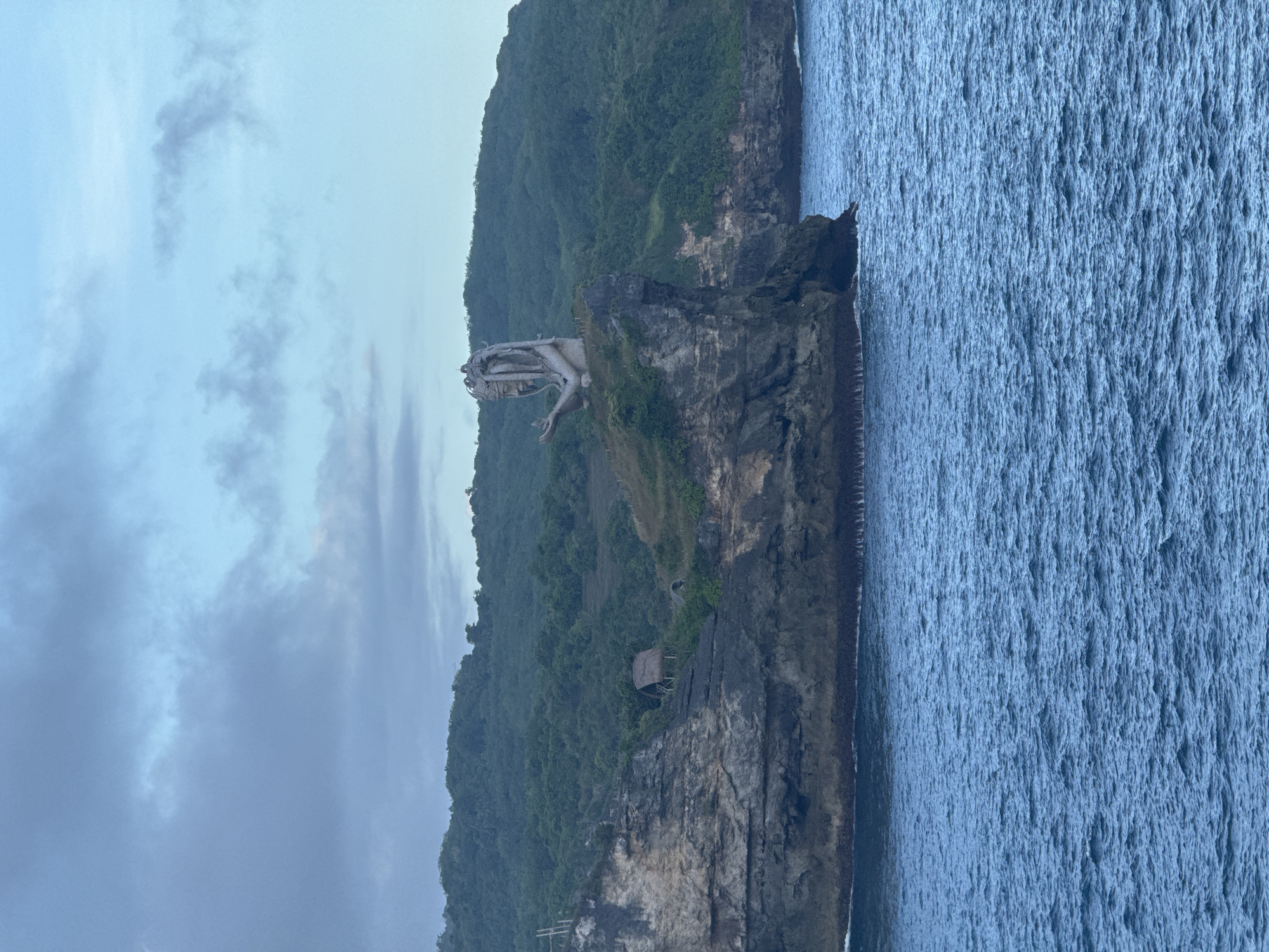 The image depicts a coastal scene in Nusa Penida, Indonesia. A large, carved statue of a mythical creature stands atop a rocky promontory that juts out into the ocean. The promontory is composed of dark, layered rock formations. The surrounding sea is a deep blue, with visible wave patterns. Lush, green vegetation covers the landscape behind the rocky outcrop. A small, wooden structure with a thatched roof is nestled on the slope below the statue. The sky is overcast with grey and white clouds, indicating possible overcast weather.