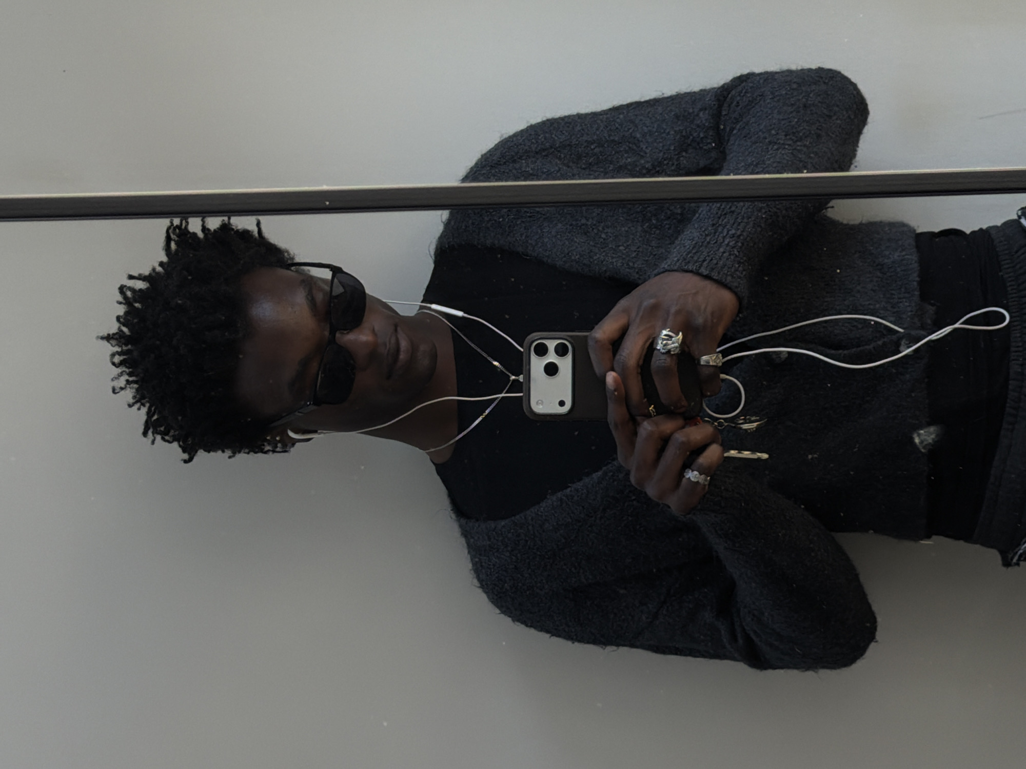 A person is depicted in a mirror selfie, taken indoors in London, United Kingdom. The image is rotated, showing the subject lying down or leaning against a surface, looking into the smartphone camera. The person has dark skin, short, curly black hair, and is wearing dark sunglasses. They are dressed in a dark grey, textured cardigan or sweater over a black top, and have white wired earphones in their ears, with the wires visible across their chest.
The person holds a dark-cased smartphone, equipped with a triple-camera system, in their right hand, using it to capture the reflection. Several rings adorn their fingers, including a distinctively shaped, possibly skull-like or abstract, ring on the right index finger. A set of keys is held in their left hand. The background is a plain, light grey wall or ceiling. A prominent dark horizontal bar stretches across the image, which could be part of the mirror's frame or a fixture within the room. No visible text is present in the image.