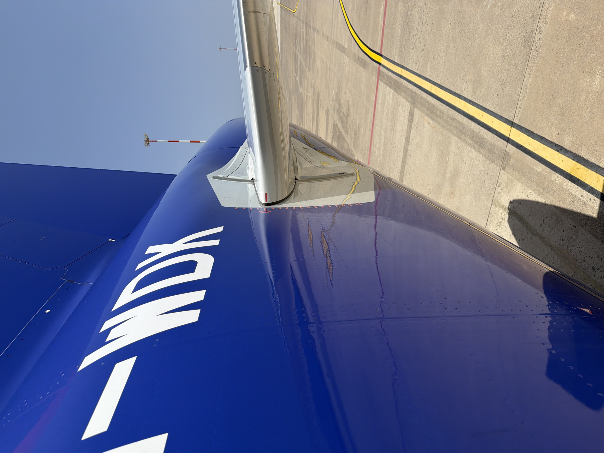 The image shows a close-up of the tail of a blue airplane with white lettering "F-WDX" on its fuselage. A portion of the wing and its mounting structure are also visible. The airplane is parked on a tarmac, indicated by the concrete ground and yellow and red painted lines. In the background, a clear blue sky is visible with a red and white striped antenna tower in the distance. The scene is outdoors, likely at an airport. The lighting suggests it is daytime. There are no people or animals visible in the image. The overall impression is one of an aircraft at rest on the ground.