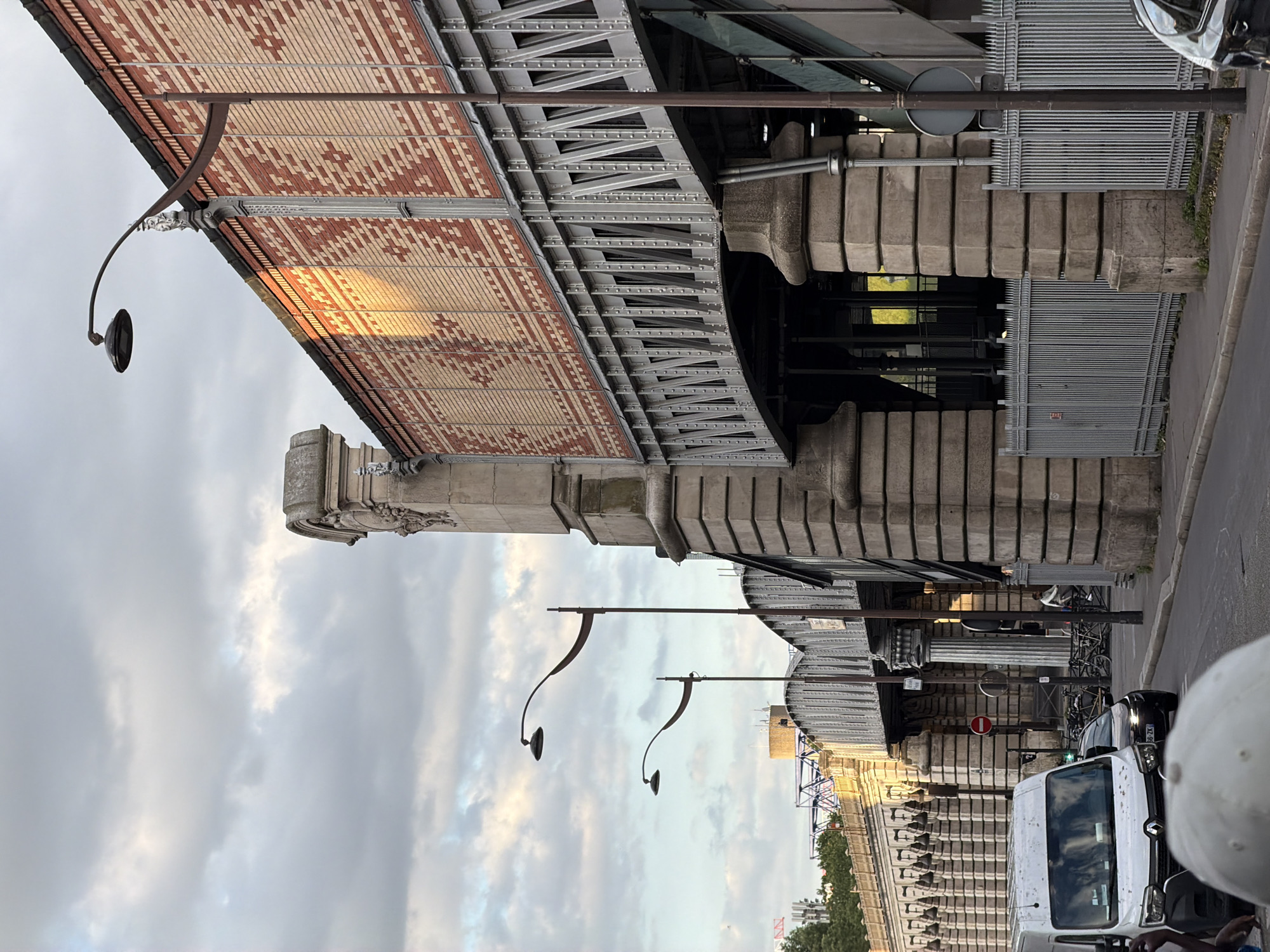 This image captures a scene in Paris, France, likely depicting a section of the Viaduc des Arts or a similar elevated structure featuring ornate brickwork and metal supports. In the foreground, a street is visible with a white van parked on the left, and a black car behind it. A person's head, wearing a white cap, is partially in the frame at the very bottom left. Further down the street, bicycles are parked, and a person is riding a bicycle on the right side of the road. The elevated structure is supported by large stone pillars, and above, a train track or walkway is visible. The architecture of the viaduct is characterized by a patterned brick facade with decorative metalwork. Streetlights with curved arms are mounted along the structure. The sky is partly cloudy, with soft sunlight casting a warm glow on the brickwork, suggesting it is late afternoon or early evening. The environment appears to be urban, with the typical infrastructure of a city.