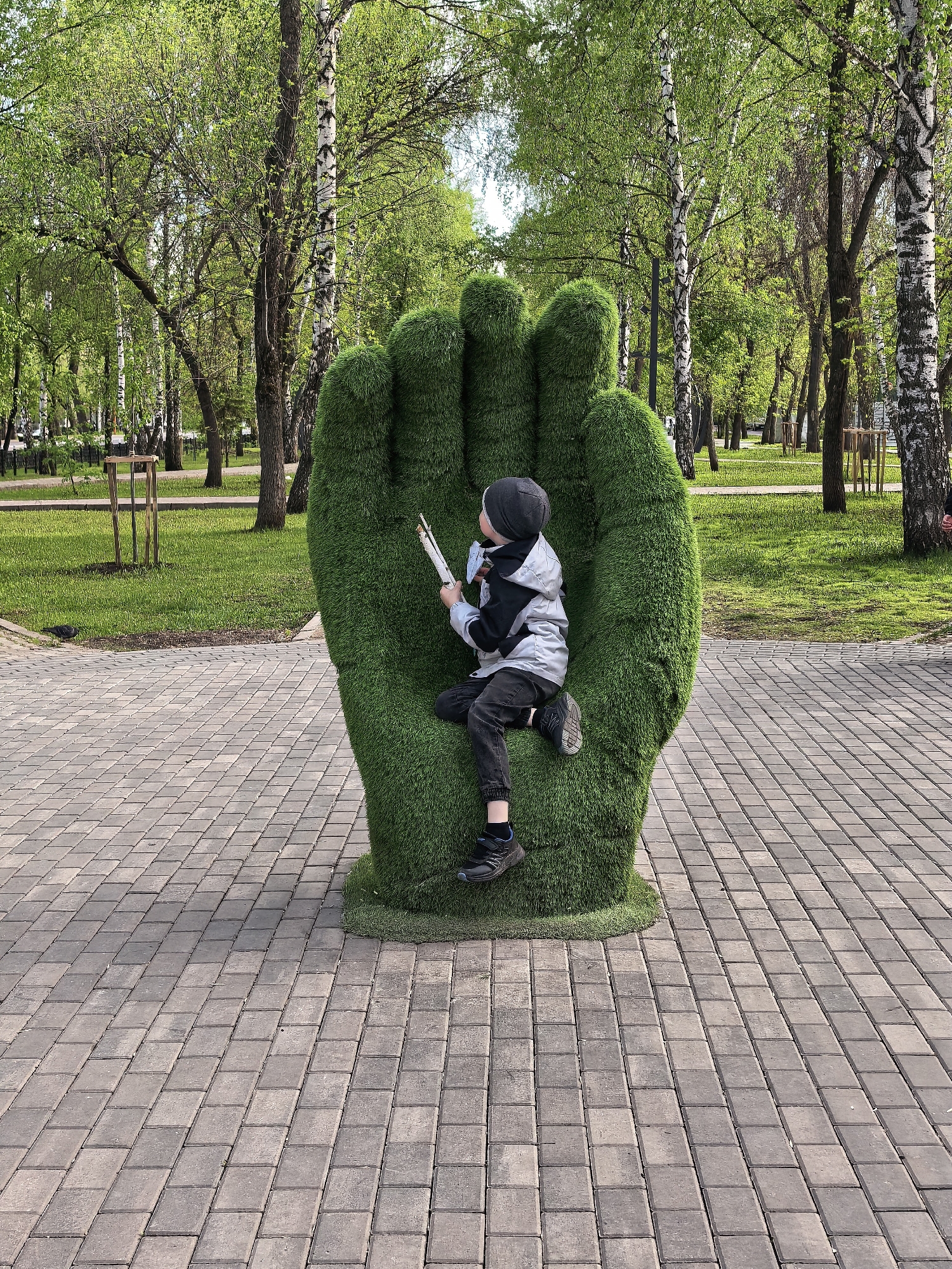 A young child sits within a large, green hand sculpture on a paved walkway in Sterlitamak, Russia. The hand sculpture is situated in a park or green space, with birch trees visible in the background. The child is wearing a dark beanie, a grey and black jacket, dark pants, and sneakers. They appear to be holding a white object. The location is Sterlitamak, Russia.