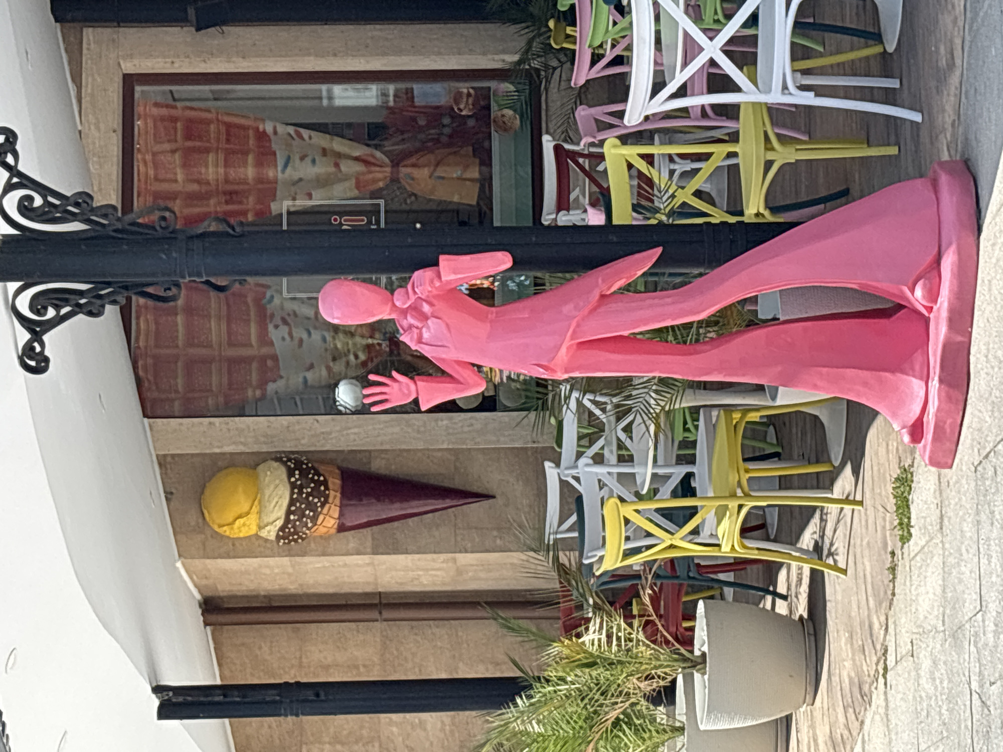 This media file depicts an outdoor scene with a focus on decorative elements outside a shop. The central subject is a large, brightly colored pink statue of a person, possibly a mannequin or a stylized sculpture, adorned in a long, flowing garment. This figure is positioned in front of a shop window displaying patterned curtains. To the lower left, a large, oversized sculpture of an ice cream cone with two scoops of ice cream sits prominently. Behind the pink statue, a collection of colorful chairs—primarily white and yellow—are stacked, suggesting an outdoor seating area for a cafe or restaurant. Palm fronds and other greenery are visible, indicating a warm climate. The ground is paved with light-colored stones. The overall impression is of a vibrant, whimsical setting, likely a commercial establishment in a tourist-oriented area. The lighting suggests it is daytime. There is no visible text in the image. The location context provided is Varna, Bulgaria.