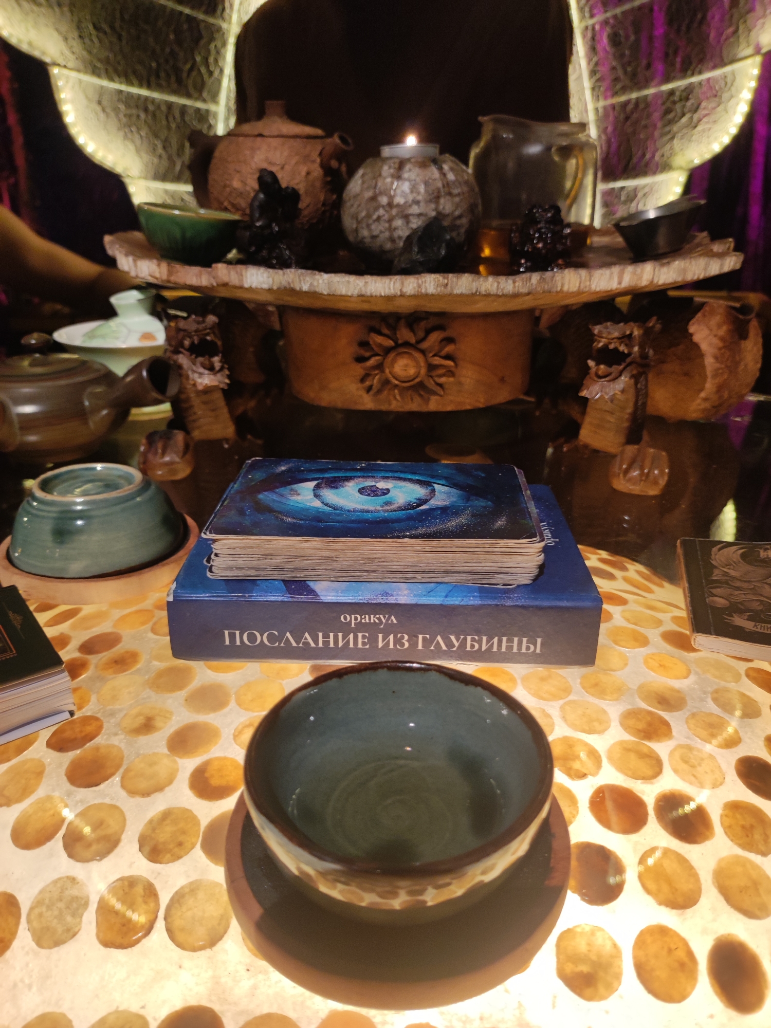 The image depicts an altar-like table, likely indoors, with various objects associated with spiritual or divinatory practices. A stack of cards with a blue eye design on top rests on a box labeled "ПОСЛАНИЕ ИЗ ГЛУБИНЫ". A teal-colored cup sits on the table in the foreground. Another cup is resting on a saucer, stacked on top of another cup, to the left of the cards. The table has a patterned surface with what appears to be round, light-colored spots. A shelf above the table holds teapots, a candle, and other small vessels. The shelf support columns are adorned with carved dragon heads. The overall setting is dimly lit and appears somewhat mystical. The location is not discernible from the image content.
