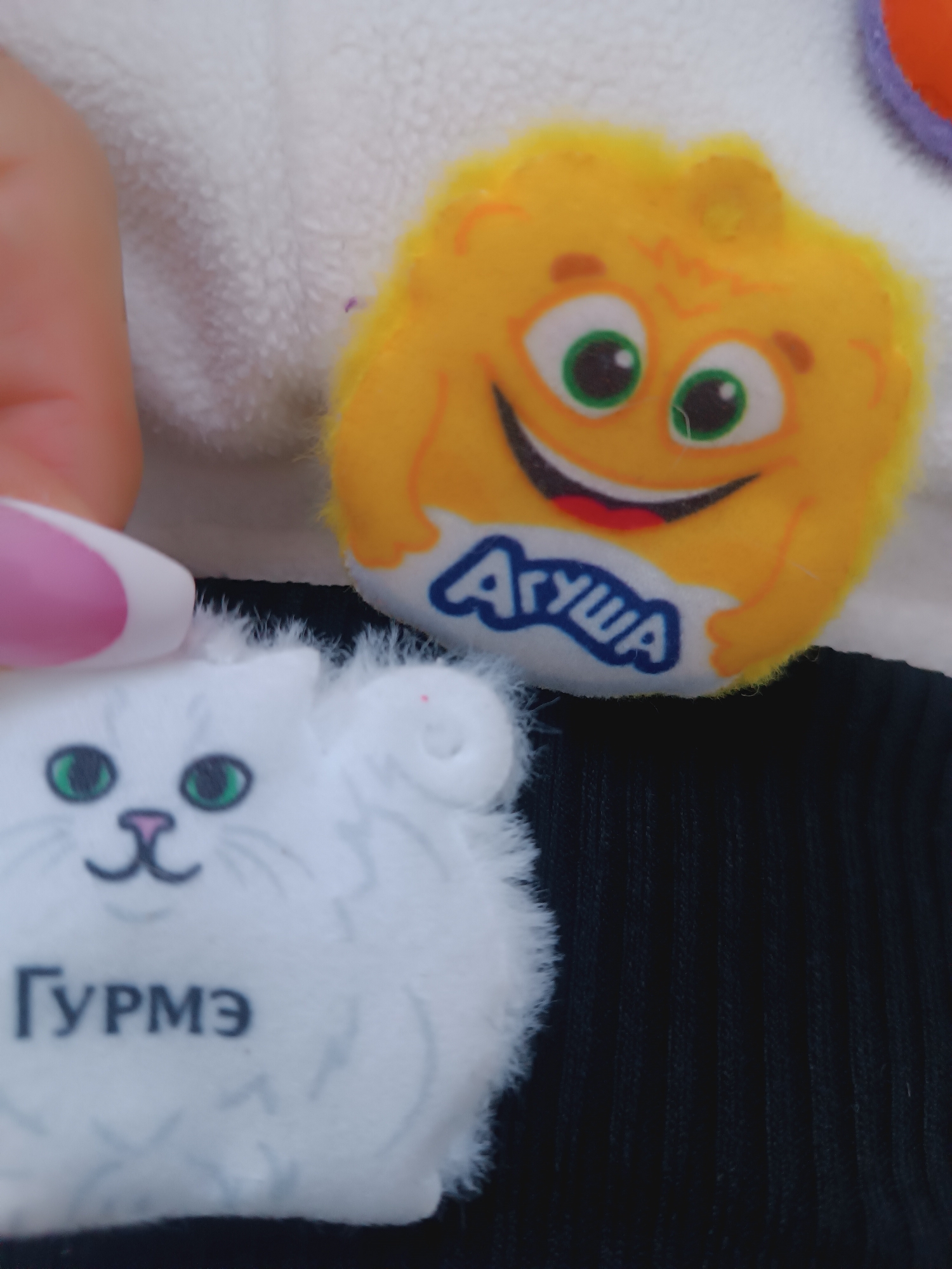 A close-up shot shows a partially visible person's finger with a pink and white nail touching a white, fluffy, cat-shaped object bearing the text "ГУРМЭ" printed on it, likely in Cyrillic. Adjacent to the cat is a yellow, round object with a smiling face and the word "АГУША" printed in blue. These objects rest against a background of black ribbed fabric and a white, fleece-like material.