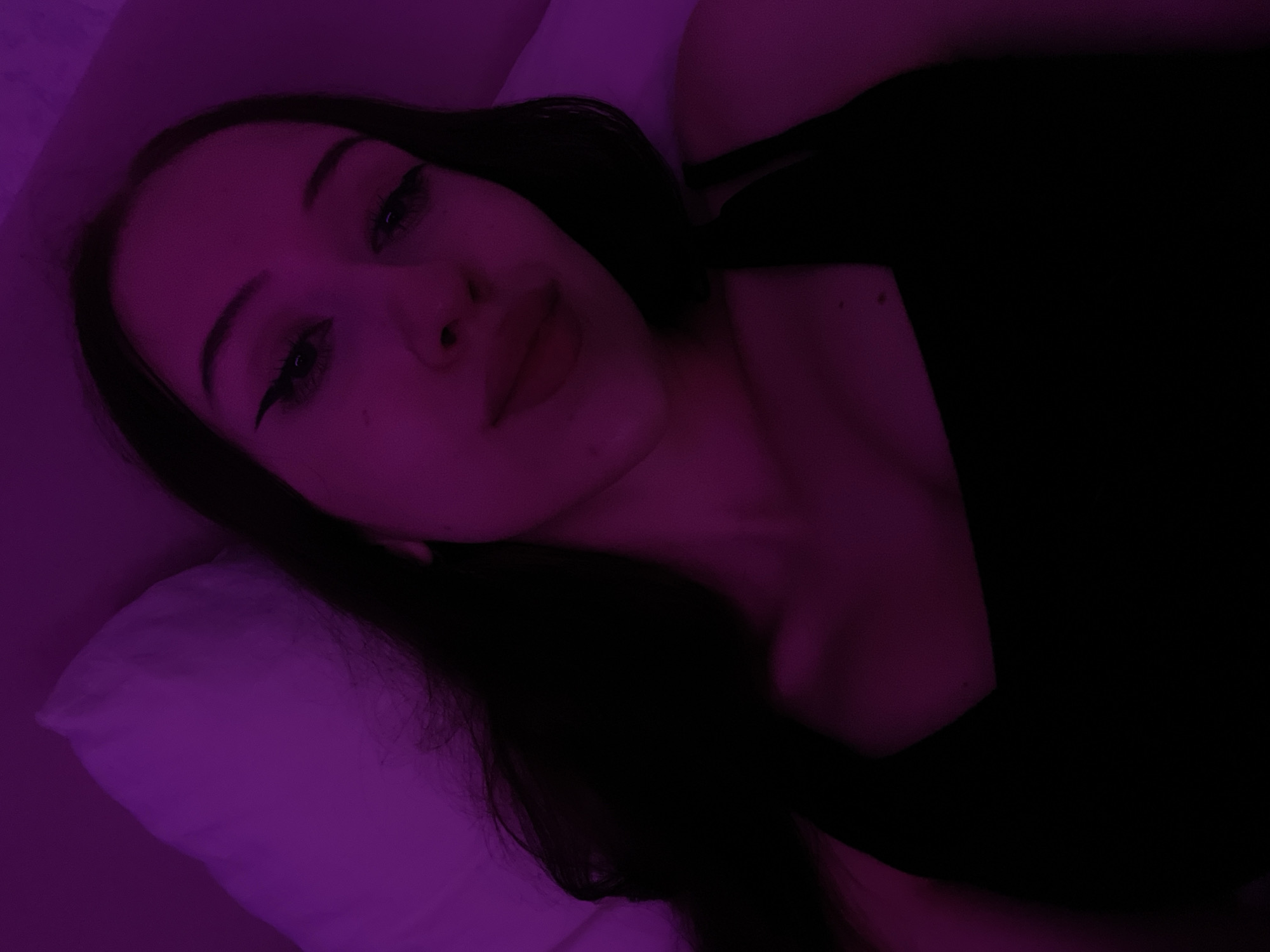 The image shows a close-up, overhead shot of a young woman lying down, possibly in bed, illuminated by purple lighting. She has long, dark hair spread around her head and shoulders. Her facial features are visible, including her eyes with winged eyeliner and her lips. She is wearing a black top. The background appears to be a pillow and possibly a wall or another surface, also lit with a purple hue. The image does not contain any discernible elements that would indicate its location in Votkinsk, Russia.