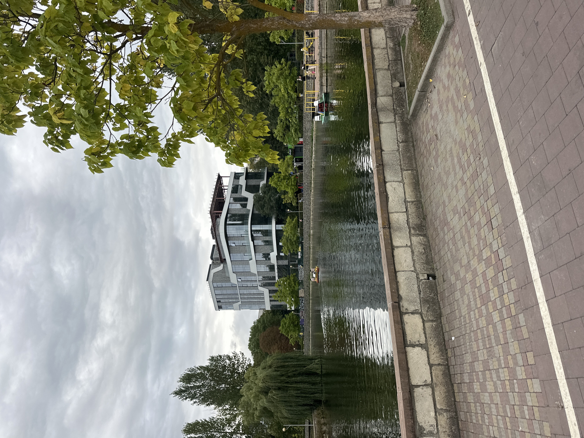 The image captures a tranquil urban park scene, likely in Ternopil, Ukraine, featuring a body of water with a modern building in the background. The overcast sky, with its soft, diffused light, contributes to the calm atmosphere. In the foreground, a red and gray brick-paved walkway runs parallel to a low, stone wall bordering a calm lake. The lake's surface reflects the surrounding greenery and the sky, with ripples indicating gentle movement.
On the lake, two paddle boats are visible. One is closer to the left side of the frame, with two people on board. Further to the right, a second paddle boat carries two individuals. Across the lake, lush green trees and shrubs form a natural backdrop, interspersed with parts of a contemporary, multi-story building characterized by its geometric design and extensive glass windows. This building appears to be an office or residential complex.
A weeping willow tree, with its characteristic drooping branches, stands prominently on the left bank of the lake, adding a touch of natural elegance. On the right side of the image, a mature tree with a textured bark stands near the walkway, its green leaves beginning to show hints of autumnal color. Along the far bank, near the building, a yellow railing delineates a pathway where several individuals can be seen walking, some with children. The overall impression is one of a well-maintained urban park, offering a space for recreation and relaxation amidst a developed cityscape.