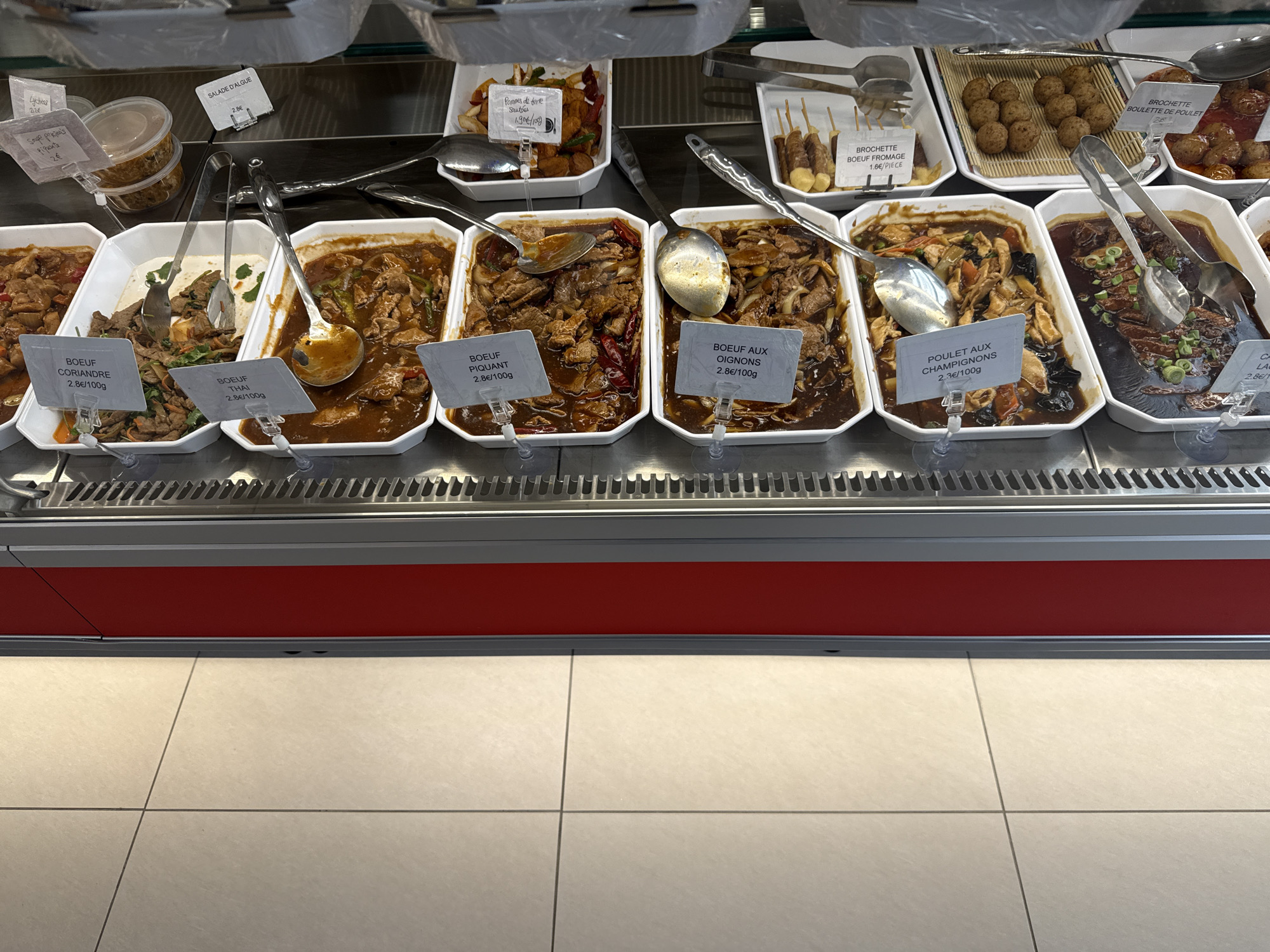 The image shows a refrigerated display case in Domont, France, filled with various prepared food items for sale. The items are arranged in white rectangular containers. The food consists mainly of beef and chicken dishes, including "Boeuf Coriandre," "Boeuf Thai," "Boeuf Piquant," "Boeuf aux Oignons," and "Poulet aux Champignons," each with a price tag of 2.8€/100g except for "Poulet aux Champignons" priced at 2.3€/100g. Additional items include skewers labeled "Brochette Boeuf Fromage," potatoes labeled "Pommes de terre Sautées," and "Brochette Boulette de Poulet." There's also a container of soup and salad. Serving spoons are placed in each of the beef and chicken dishes. The background shows more food items, including what appear to be meatballs and other brochettes. The floor in front of the display case is tiled.