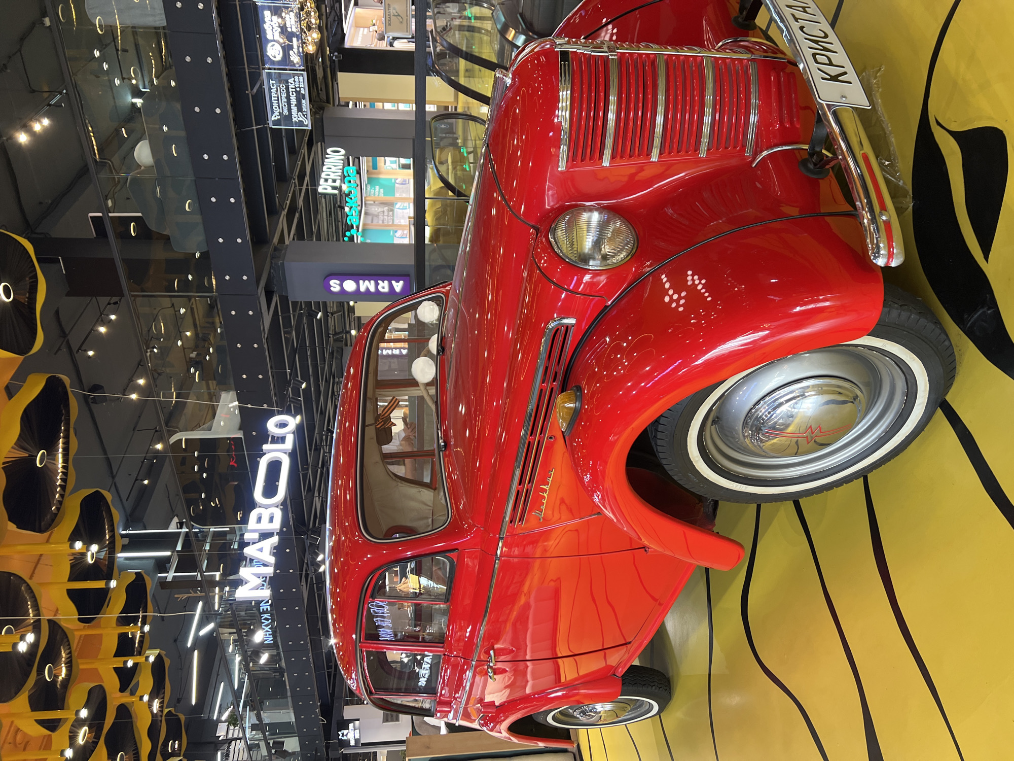 The image captures a vintage red car, possibly a Moskvich, prominently displayed in what appears to be a shopping mall or a public space in Moscow, Russia. The car, which has a license plate reading "КРИСТА" (KRISTA), is parked on a yellow surface with black abstract patterns.
In the background, there are various commercial signs, including "MAIBOLO", "ARMOS," and "PERRINO," indicating the presence of different stores or brands. Additional signs in Russian, such as "КОНТРАСТ ЭКСПРЕСС ХИМЧИСТКА," which translates to "CONTRAST EXPRESS DRY CLEANING," further confirm the location.
The environment is a modern shopping mall with glass panels, escalators, and decorative lighting fixtures, creating a bustling and vibrant atmosphere. The image seems to be taken from a slightly low angle, emphasizing the height and layout of the mall.