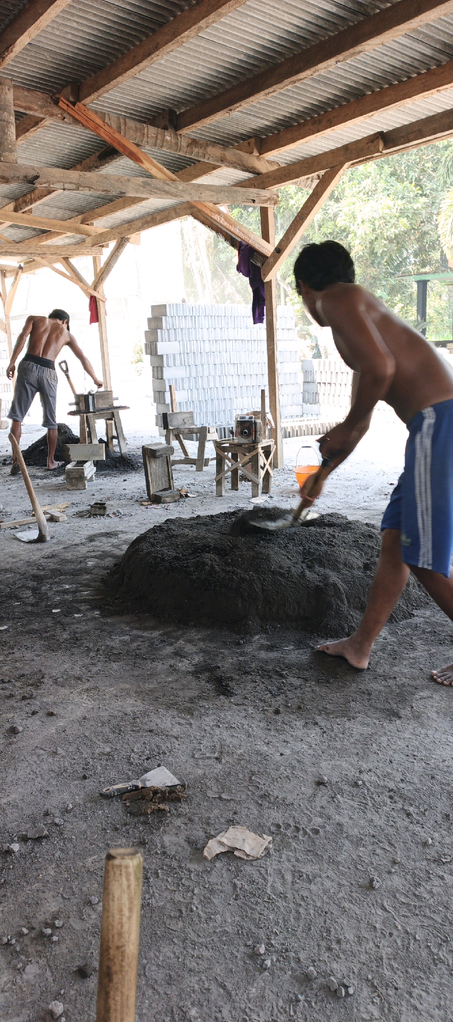 Two shirtless men are working on a construction site under a corrugated metal roof supported by wooden beams. The ground is dirt and gravel. In the foreground, a large pile of dark, wet cement is being mixed by a man using a shovel. He is wearing blue shorts with white stripes. In the background, another man is operating a cement brick mold with a shovel. He is wearing gray shorts. Behind him are stacks of white cement bricks. There are various wooden structures and tools scattered around the site. The scene appears to be in Kasiyan, Indonesia.