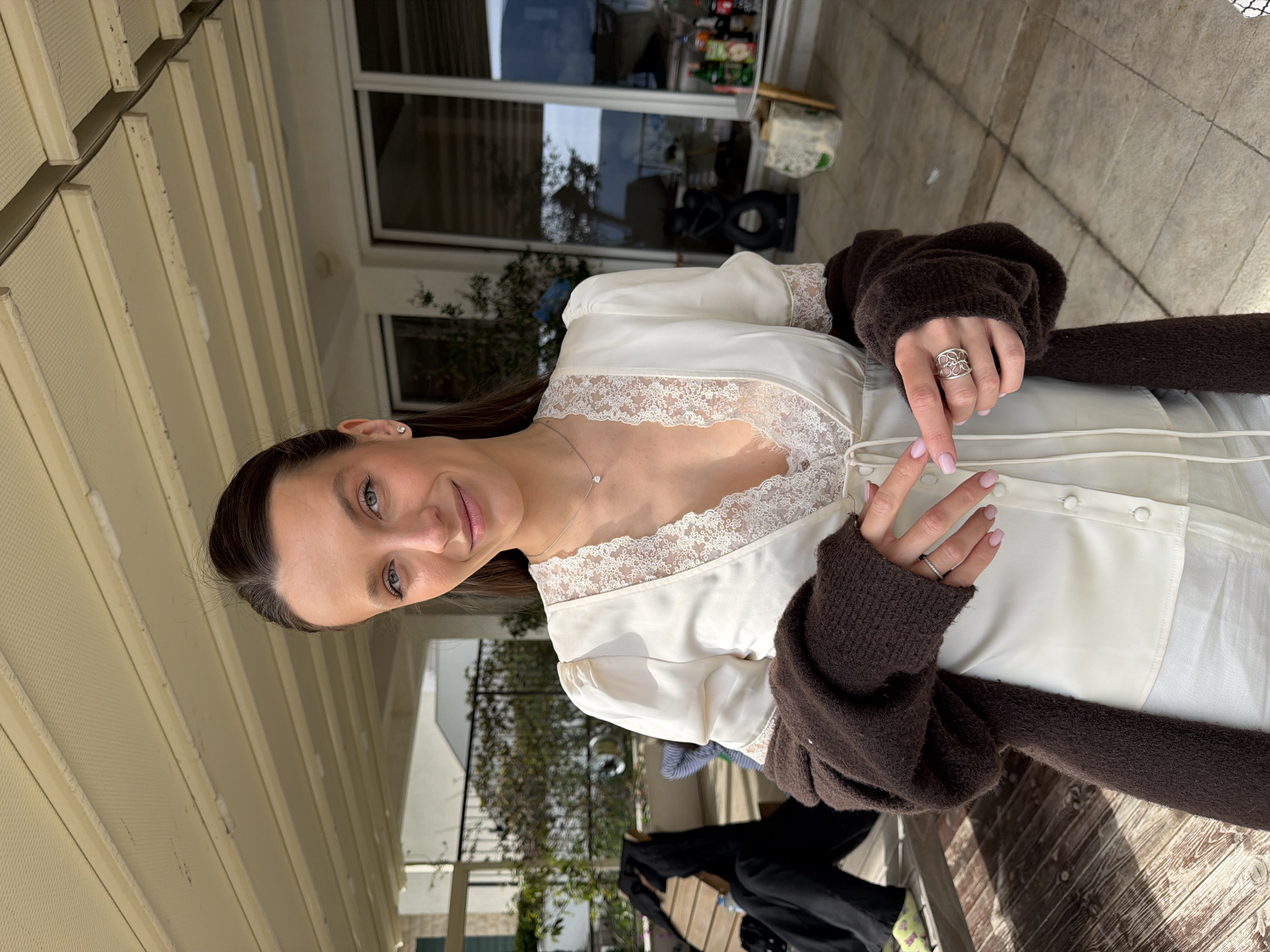 A waist-up portrait of a woman with dark hair and fair skin. She is wearing a white, lacy top under a brown sweater. She is holding a white drawstring, possibly to tie her top. The photo is taken outside, with the woman standing on a patio. In the background, there is a house with large glass windows and greenery. The setting appears to be in the city of Trebesin, Montenegro.