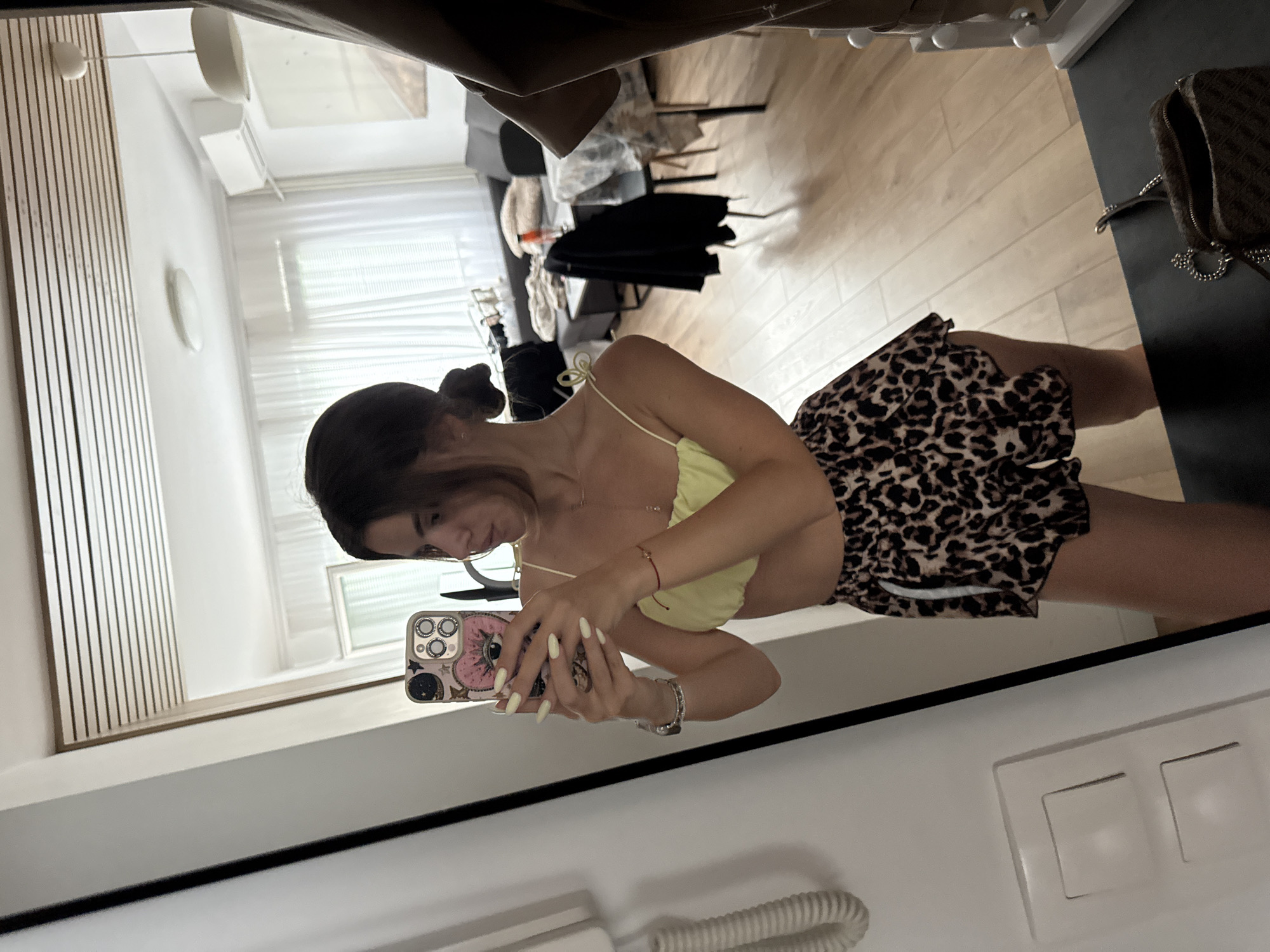 A young woman with dark hair pulled back in a bun is taking a mirror selfie in what appears to be a modern apartment. She is wearing a light yellow spaghetti strap top and leopard-print shorts. She is holding her phone, which has a decorative case featuring a pink heart and eye design, in her right hand. Her left hand is resting on her hip. A silver bracelet is visible on her left wrist. In the reflection of the mirror, a living area can be seen with a sofa, a coffee table with items on it, and sheer curtains covering a window. A jacket is hanging on a coat rack to the right of the woman. The floor is made of light-colored wood planks. The overall scene suggests a casual moment captured in a residential setting, likely an apartment in Plovdiv, Bulgaria.