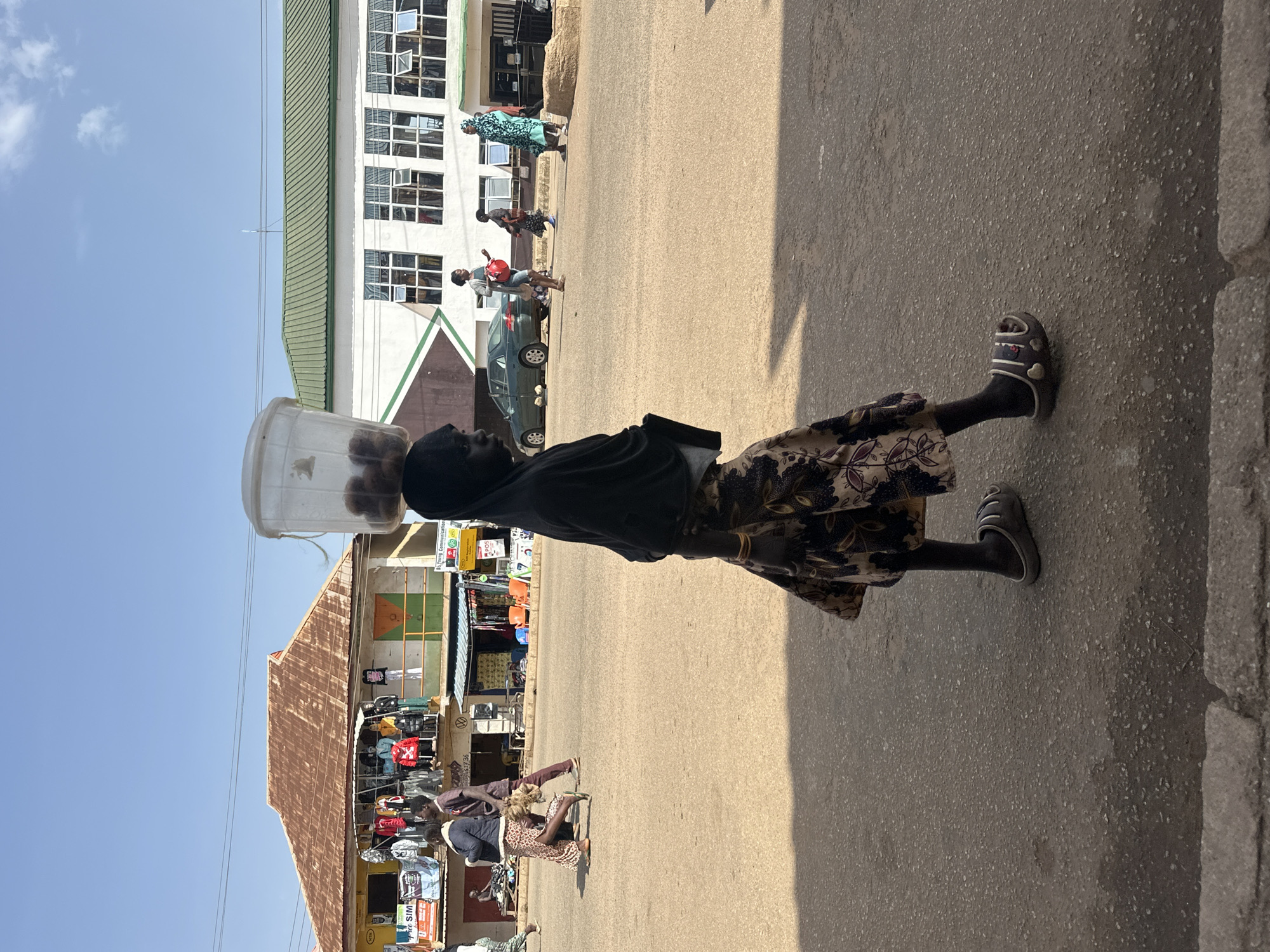 In the city of Bukuru, Nigeria, a young girl is captured in a side profile walking across a dusty, paved road. She wears a dark headscarf and a floral print skirt. Perched precariously on her head is a clear plastic bucket filled with what appear to be round, brown fruits, possibly yams or potatoes. Her feet are clad in dark, open-toed sandals. In the background, a row of low-rise buildings lines the street. The building on the left has a weathered, reddish-brown corrugated metal roof and displays various clothing items on racks outside its ground floor. The building on the right is a more modern, white structure with a green corrugated metal roof and multiple windows. Other individuals are visible walking or standing in the mid-ground. A green sedan is parked on the road near the white building. The sky above is clear blue.