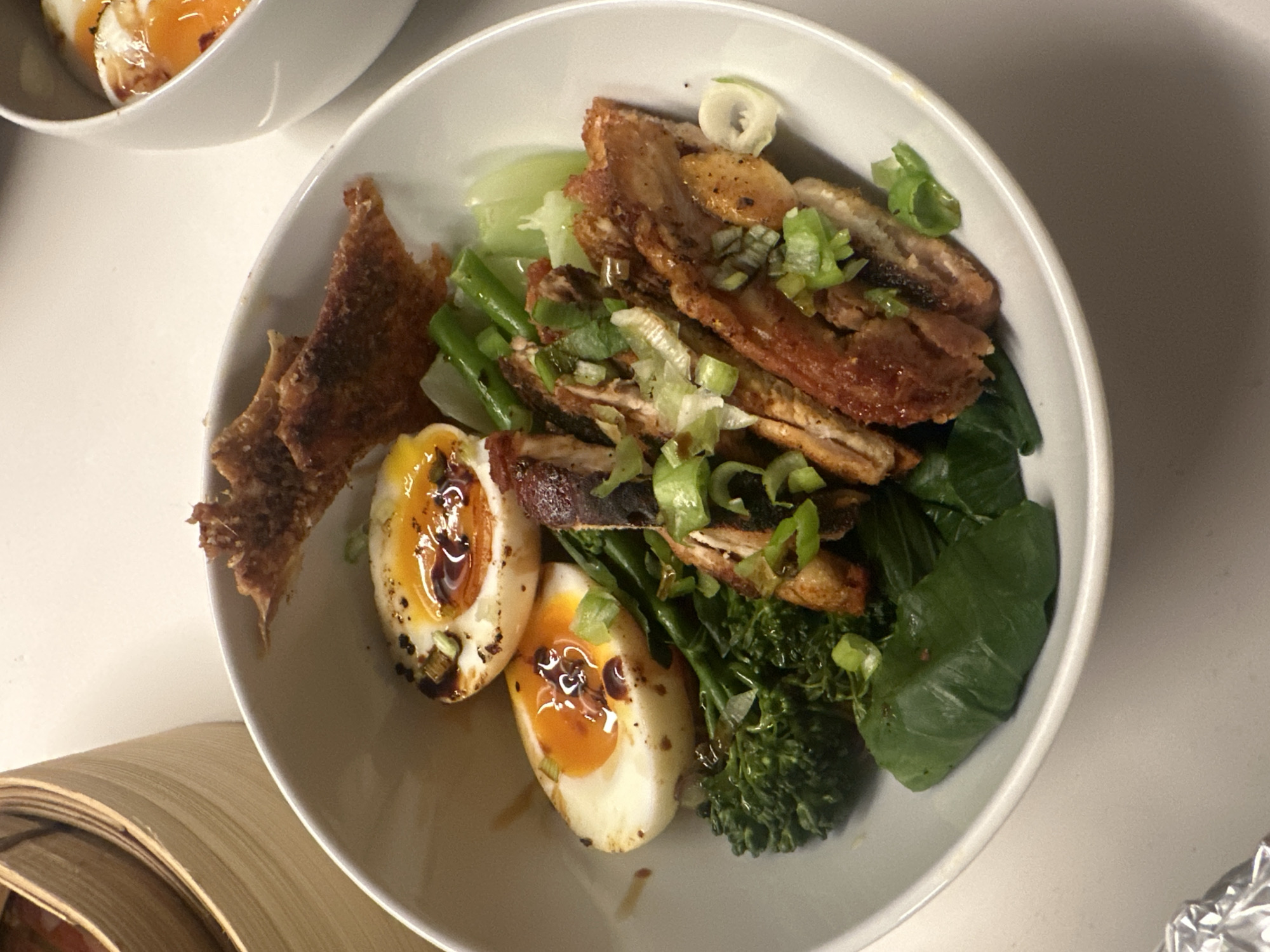 A close-up, eye-level shot shows a white bowl filled with food. The bowl contains sliced meat (possibly pork belly) topped with chopped green onions, alongside grilled bok choy, stir-fried green beans, two grilled eggs with runny yolks drizzled with sauce, and what appears to be fried wonton skins. The ingredients suggest an Asian-inspired dish. The bowl is placed on a white surface, with the edge of another bowl visible in the upper left corner and a partially visible bamboo steamer in the lower left. The image does not provide information about the location of the photo.
