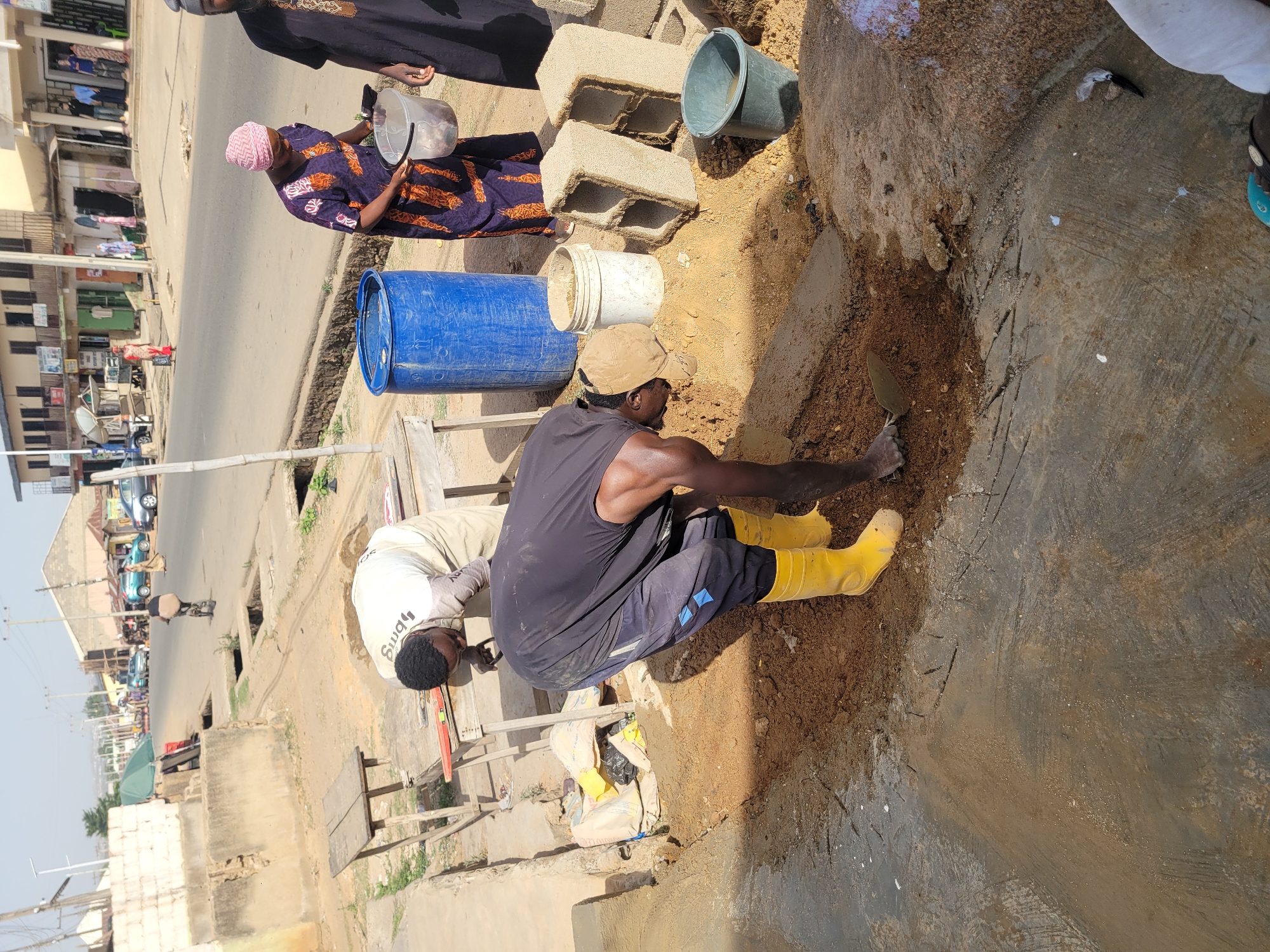 This image captures an outdoor construction or repair scene in Jos, Nigeria, under bright daylight.
In the foreground, a man wearing a black tank top, dark pants, and yellow rubber boots is kneeling in a dug-out earthen trench, actively working with a trowel. Next to him, another man in a white t-shirt with "BBMG" visible on the back, and light-colored pants, is bending over a makeshift wooden structure, seemingly engaged in a related task. Behind these men, a woman dressed in a patterned purple and orange/brown dress and a pink headscarf stands, holding a clear plastic container. Another individual in dark clothing is partially visible at the top edge of the frame.
Various construction materials are scattered around the site, including hollow concrete blocks, a blue plastic barrel, a white bucket, and a grey/green bucket. Bags, possibly containing cement or sand, are also visible. A red and clear level tool rests on the wooden structure. The ground consists of both excavated earth and a concrete surface.
The background reveals a bustling street scene with several multi-story buildings, cars, and a person riding a bicycle, typical of an urban or semi-urban environment. The strong shadows indicate clear, sunny weather, likely in the mid-morning or afternoon. The overall impression is one of manual labor being carried out in a public space.