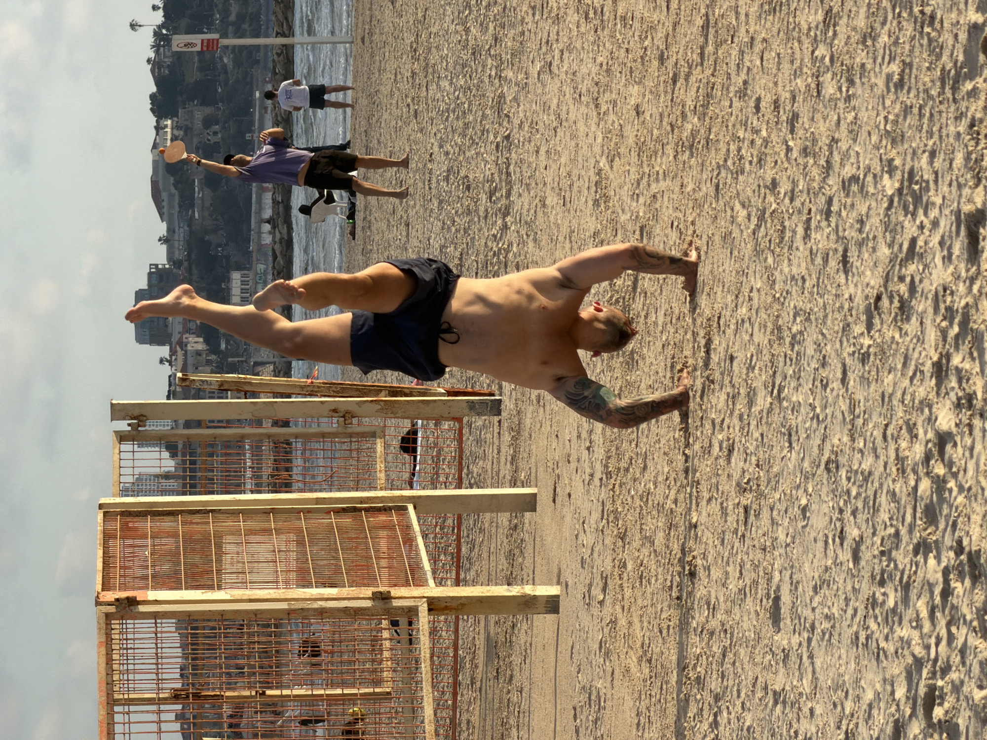 On a sunlit beach in Tel Aviv, Israel, a muscular man with tattoos performs a handstand on the sand. He wears dark blue shorts. In the background, other people are playing matkot (paddleball). Beyond them, the Mediterranean Sea meets the city's skyline. An old rusted metal staircase is situated to the left of the man. The overall scene is captured in a clear, bright, outdoor setting.