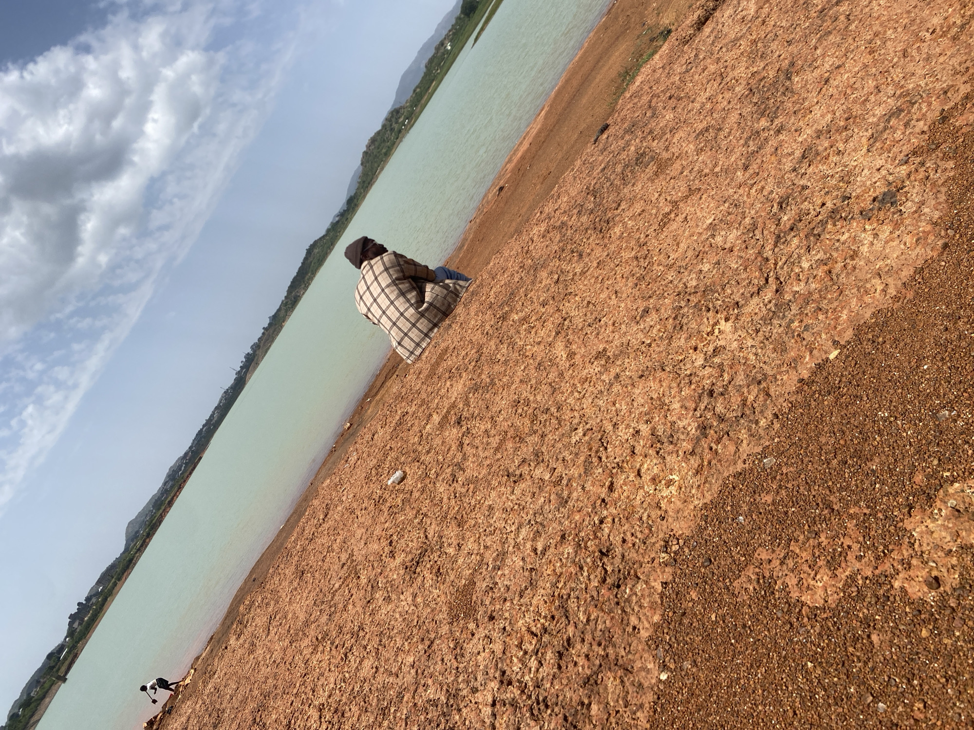 A wide-angle shot shows a person sitting by the water's edge in Jos, Nigeria. The person is wearing a checkered shirt and a dark cap, facing away from the camera. The water body is pale, bordered by a reddish-brown ground with varied texture. In the background, a land mass is visible. The sky overhead is partly cloudy. At the far left corner is another person walking along the shoreline.