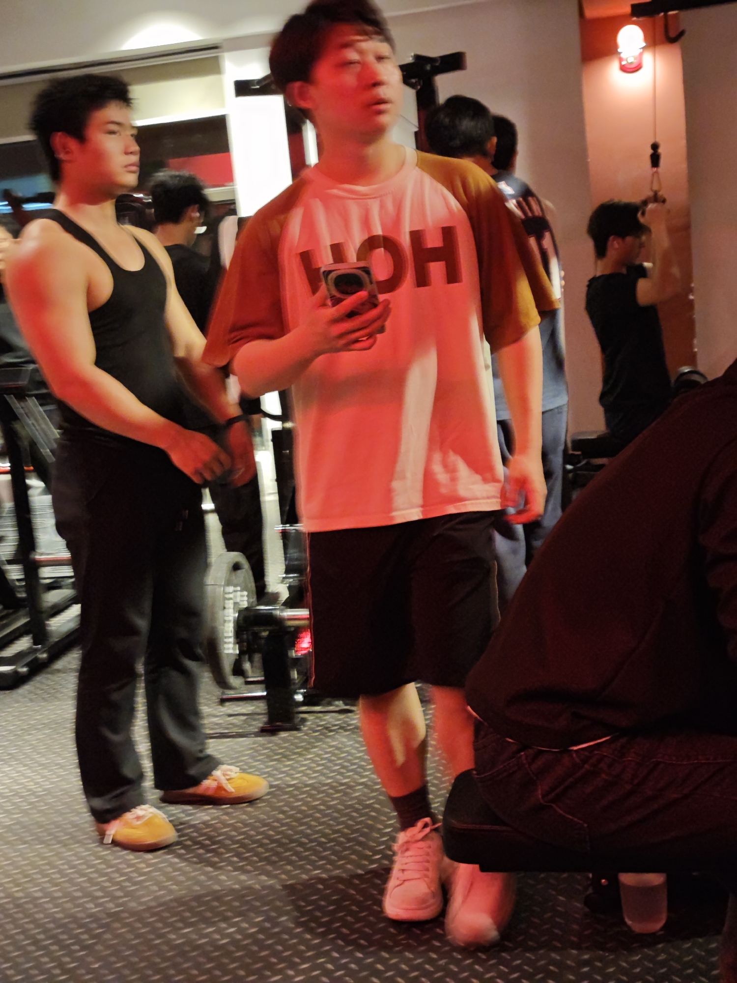Several people are in a gym. A man in a black tank top and dark pants with yellow sneakers stands near a treadmill. Another man in a white shirt with brown sleeves, black shorts, and pink sneakers is holding a phone. In the background, another man is using a pull-down machine. The floor has a diamond pattern. The gym lighting is red and white.