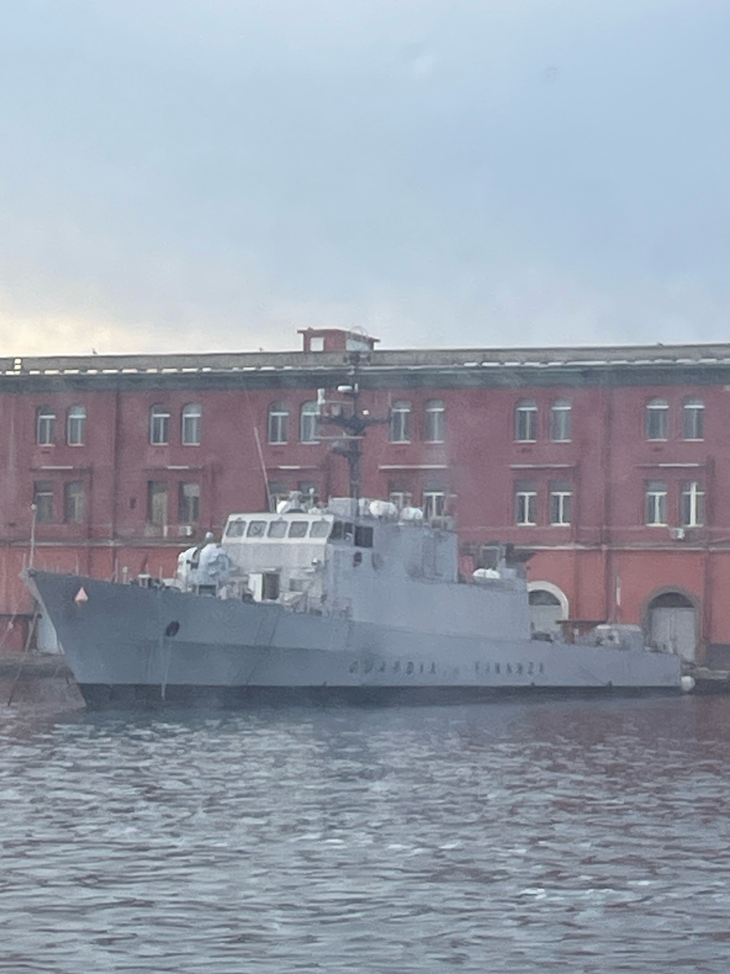 A gray patrol boat with the words "GUARDIA FINANZA" visible on its side is docked in front of a large, red brick building in Naples, Italy. The building has multiple arched doorways and windows. The sky is overcast, and the water appears calm. The boat is the primary subject, with the building serving as a backdrop. The scene is static and appears to be taken from a distance.