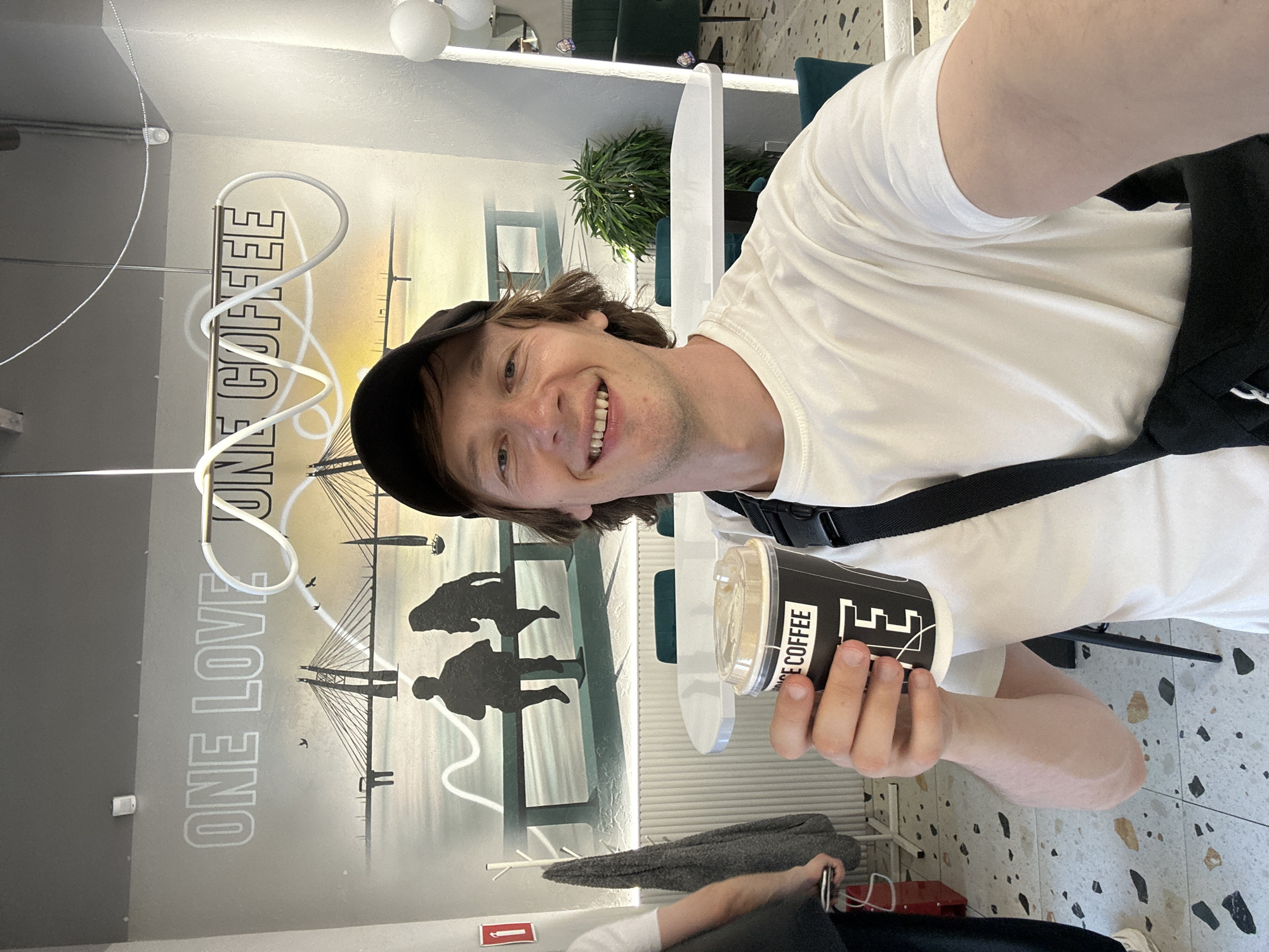 A man with brown hair and a black baseball cap is taking a selfie in what appears to be a cafe. He is wearing a white t-shirt and a black strap across his chest. He is smiling and holding a black paper cup with a clear lid. The cup has the words "ONE LOVE COFFEE" printed on it in white text.
Behind the man, there is a mural on the wall. The mural features the text "ONE LOVE COFFEE" in large, stylized white letters. Below this text is a silhouette of a bridge, reminiscent of the bridges in Saint Petersburg, Russia, with two silhouetted figures standing in the foreground. The sky in the mural is a gradient of yellow and white, suggesting a sunrise or sunset.
The cafe has a modern interior with speckled terrazzo flooring and teal chairs visible in the background. There are also some hanging white spherical lights and a green plant. The overall impression is that of a casual, bright, and contemporary setting, likely during the daytime. The man's expression conveys happiness and a relaxed mood.