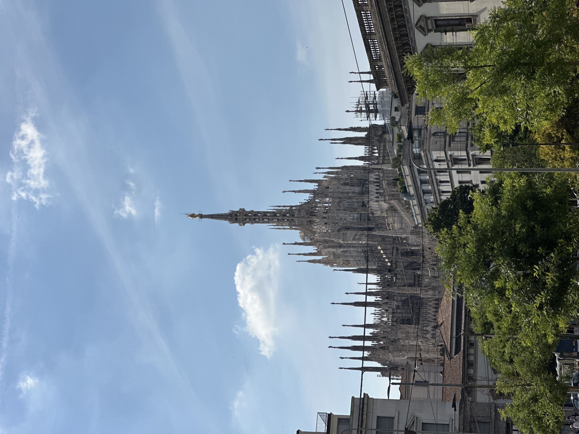 This forensic description details an outdoor scene dominated by the Milan Cathedral (Duomo di Milano) in Milan, Italy, viewed from a slightly elevated urban perspective under a bright, partly cloudy sky.
The Milan Cathedral occupies the central and upper-mid portions of the image, showcasing its intricate white marble Gothic architecture with numerous spires. The tallest central spire, capped by the golden Madonnina statue, is prominently visible against the sky. Multiple other spires, each adorned with a statue, are visible across the cathedral's roofline.
The sky above is clear blue with scattered white clouds, including a distinct cumulus cloud positioned to the left of the main spire and faint wispy clouds and contrails higher up.
In the foreground and mid-ground, various urban elements frame the cathedral. To the left, a multi-story building with a light-colored facade and a rooftop balcony partially obscures the cathedral's lower left side. To the right, an ornate building with a decorative rooftop balustrade is visible. Between these, several other buildings, including those with terracotta tiled roofs, are discernible. Scaffolding is erected around a structure or section of the cathedral complex visible to the right of the main spires.
Numerous utility poles and overhead wires crisscross the mid-ground horizontally, partially obscuring the cathedral's lower sections. The bottom third of the image is filled with the lush green foliage of a row of deciduous trees, further obscuring the street level. Indistinct human figures are visible through the trees at the bottom left, and a portion of a blue vehicle, possibly a bus or tram, is faintly visible through the trees at the bottom center. The scene is uniformly illuminated by natural daylight, indicating a sunny period.