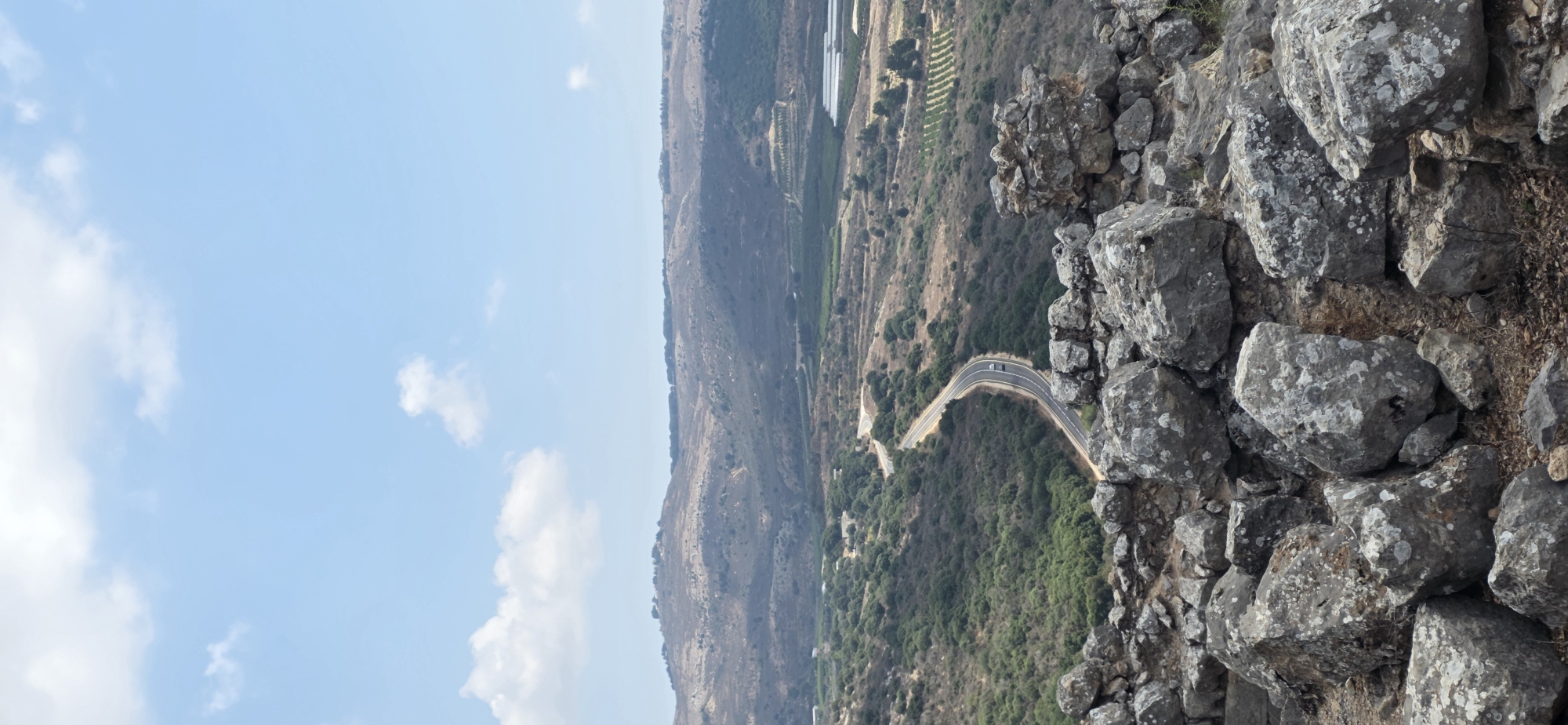 The image captures a scenic view of the Golan Heights region in Israel. In the foreground, a pile of weathered grey rocks forms a natural barrier, suggesting an elevated viewpoint. Beyond the rocks, a winding asphalt road snakes through a verdant valley, with cars visible on the road. The valley is dotted with dense green foliage and patches of dry, arid land, indicative of a Mediterranean climate. In the distance, rolling hills stretch towards the horizon, their slopes covered in sparse vegetation and some settlements. The sky above is a pale blue, with scattered white clouds, suggesting a clear, sunny day. The overall scene conveys a sense of natural beauty and a rural landscape.
