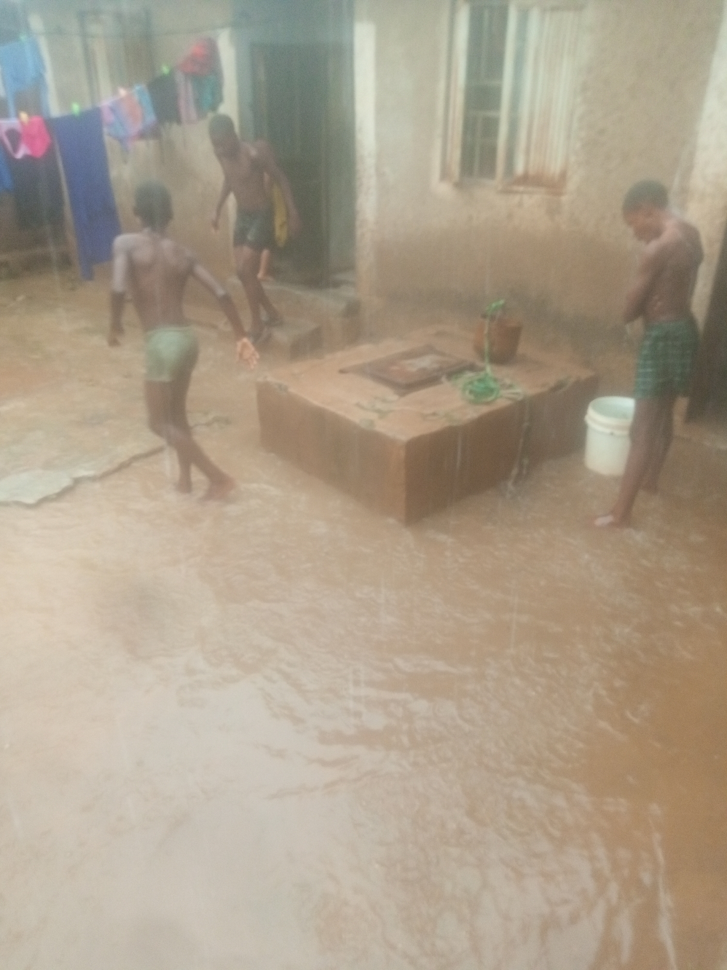 The image depicts an outdoor scene in Unknown, Unknown, under heavy rainfall. Three shirtless boys are wading through floodwater. One boy is moving to the left side of the frame with outstretched arms. A second boy is entering a doorway on the left side of the frame. The third boy stands on the right side of the frame, seemingly in the rain. A square, concrete structure is present in the center of the image, with a bucket, rope, and cover on top. In the background, clothing hangs on a clothesline, and a building with a window is visible. The ground is covered in muddy water due to the heavy rain.