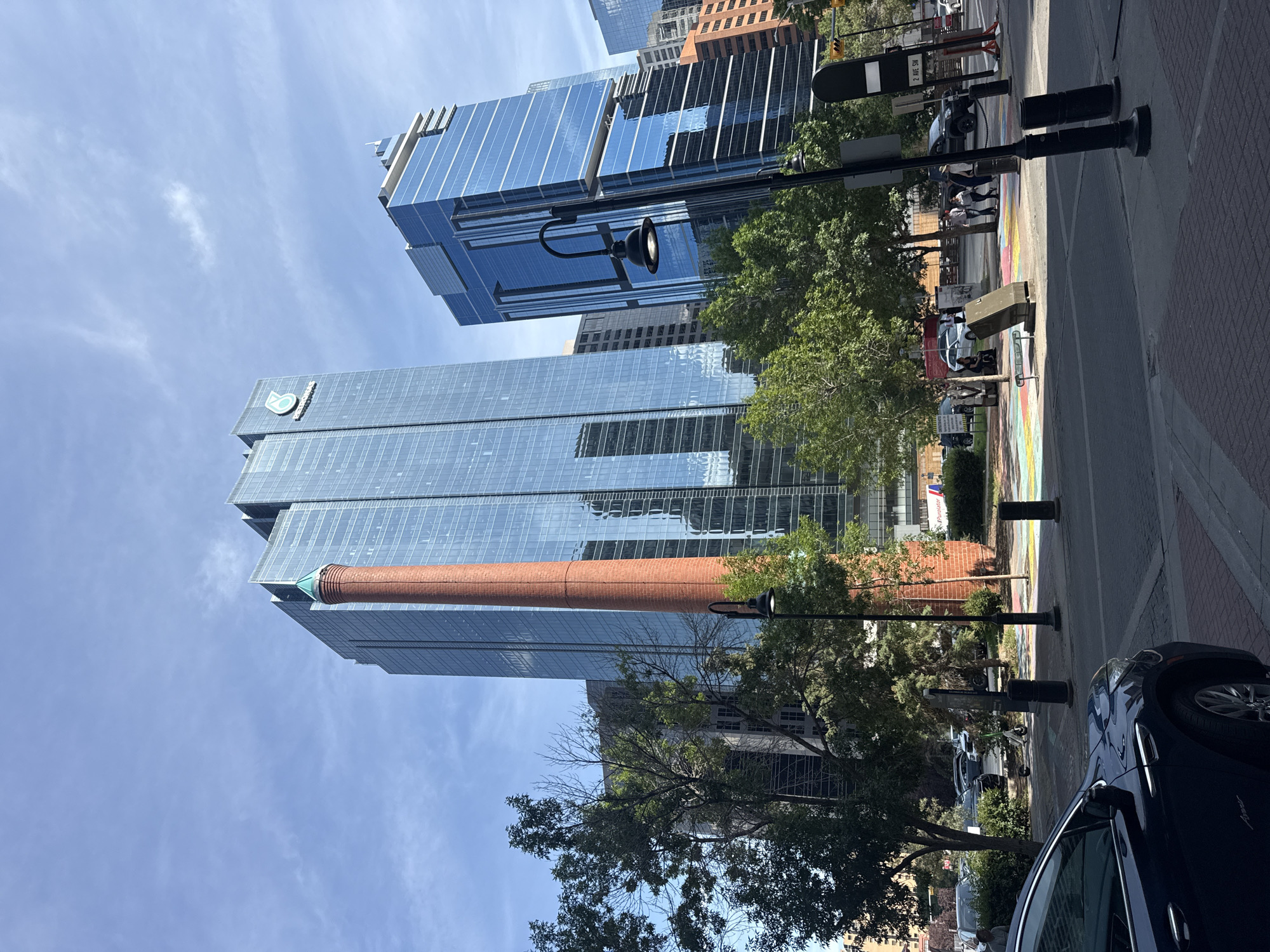 The image shows the Petro-Canada building and the adjacent Encor Tower in downtown Calgary, Canada, with a prominent brick smokestack in the foreground. The sky is blue with some scattered clouds. The Petro-Canada building is a tall, modern skyscraper with a glass facade, and the Encor Tower is to its right, also a glass-fronted building. The brick smokestack, a historical remnant, stands tall next to the modern buildings. Trees with green foliage are visible in front of the buildings, and a street with cars, people walking, and traffic signals occupies the lower part of the image. There is also a colorful mural painted on the sidewalk. The scene appears to be a sunny day.