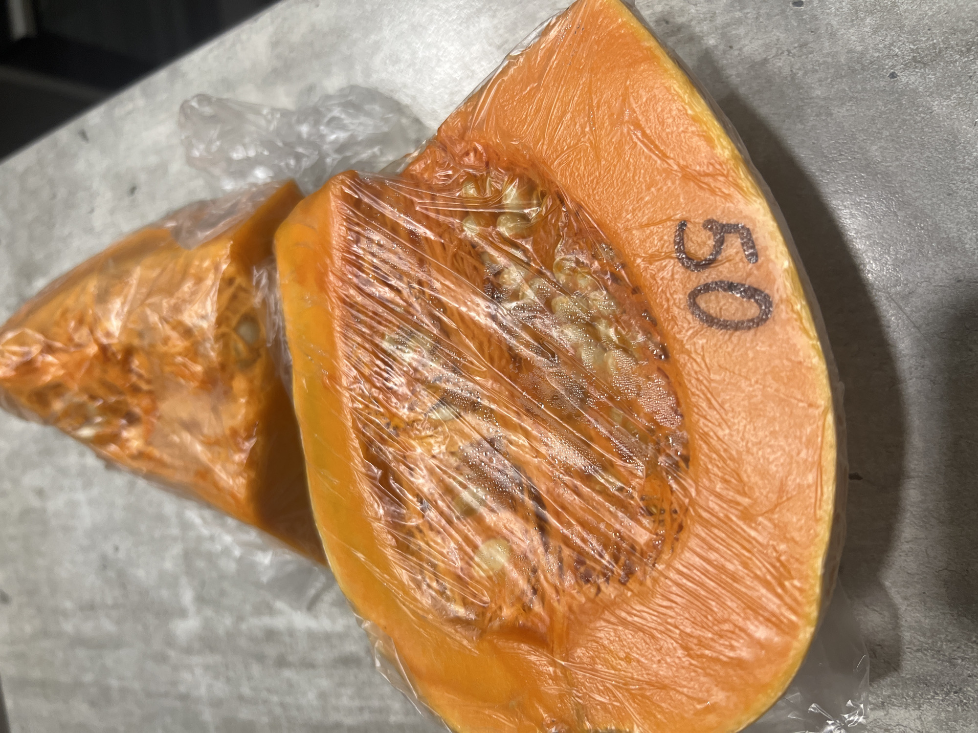 Two slices of orange pumpkin, wrapped in clear plastic, are displayed on a grey, textured surface. One slice is in the foreground, with a segment of the pumpkin's seeds and fibrous interior visible through the plastic. The number "50" is written in dark ink on the rind of this slice. A second slice is partially visible behind the first, also wrapped in plastic. The lighting suggests an indoor setting, possibly a kitchen or market. The image appears to be a close-up shot, focusing on the details of the pumpkin slices. There are no people or animals present.
