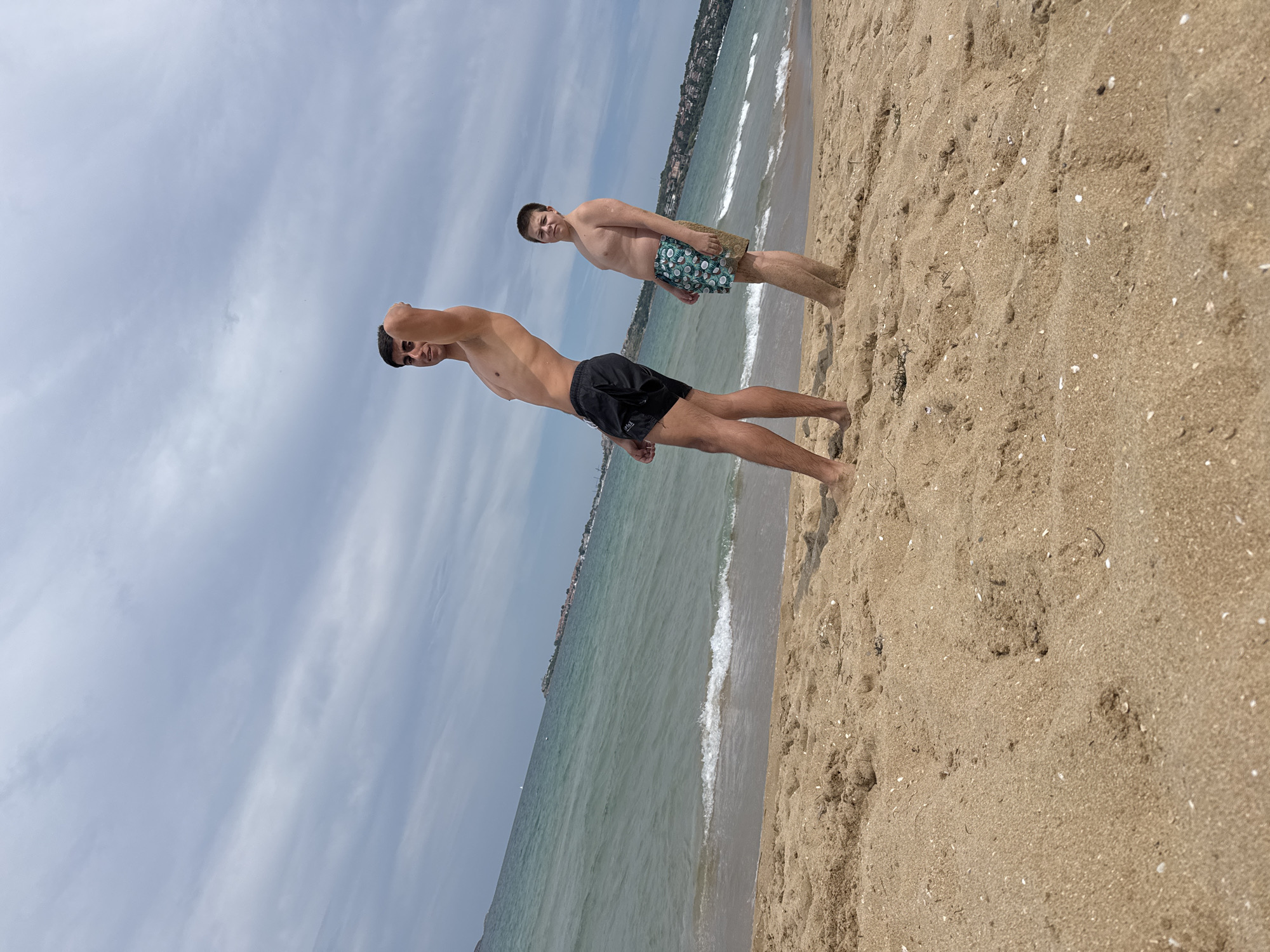 This is a low-angle, full shot of two young males standing on a sandy beach near Sozopol, Bulgaria. The sky is cloudy and overcast. In the foreground, the sand is textured with small shells and debris. The sea is visible in the middle ground, with gentle waves lapping at the shore. In the background, there are buildings and vegetation on a coastline, suggesting the presence of a town or resort.
The male on the left is taller and leaner, with dark hair and a tan. He is shirtless, wearing black swim trunks, and poses with one arm raised, hand on his head. The male on the right is shorter and heavier-set, with light brown hair. He is also shirtless, wearing patterned swim trunks. He stands with his arms down at his sides, looking towards the camera with a neutral expression. Both individuals appear to be on a beach vacation.