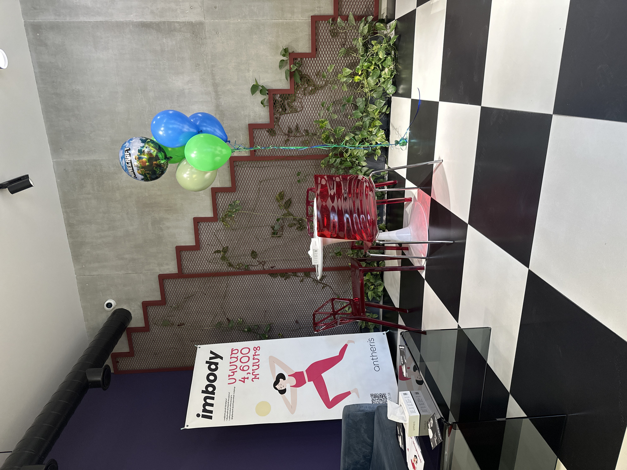The image shows an interior setting with a black and white checkered floor, a gray wall with red stair-step trim, and climbing plants against the wall. A collection of balloons in blue, green, and a Minecraft-themed design are present. There's a red translucent chair and a banner that reads "Imbody" in Armenian with an illustration of a woman in a yoga pose, along with the word "ontheris". The objects and text suggest a fitness or wellness context. The location is Yerevan, Armenia, based on the Armenian language on the banner.