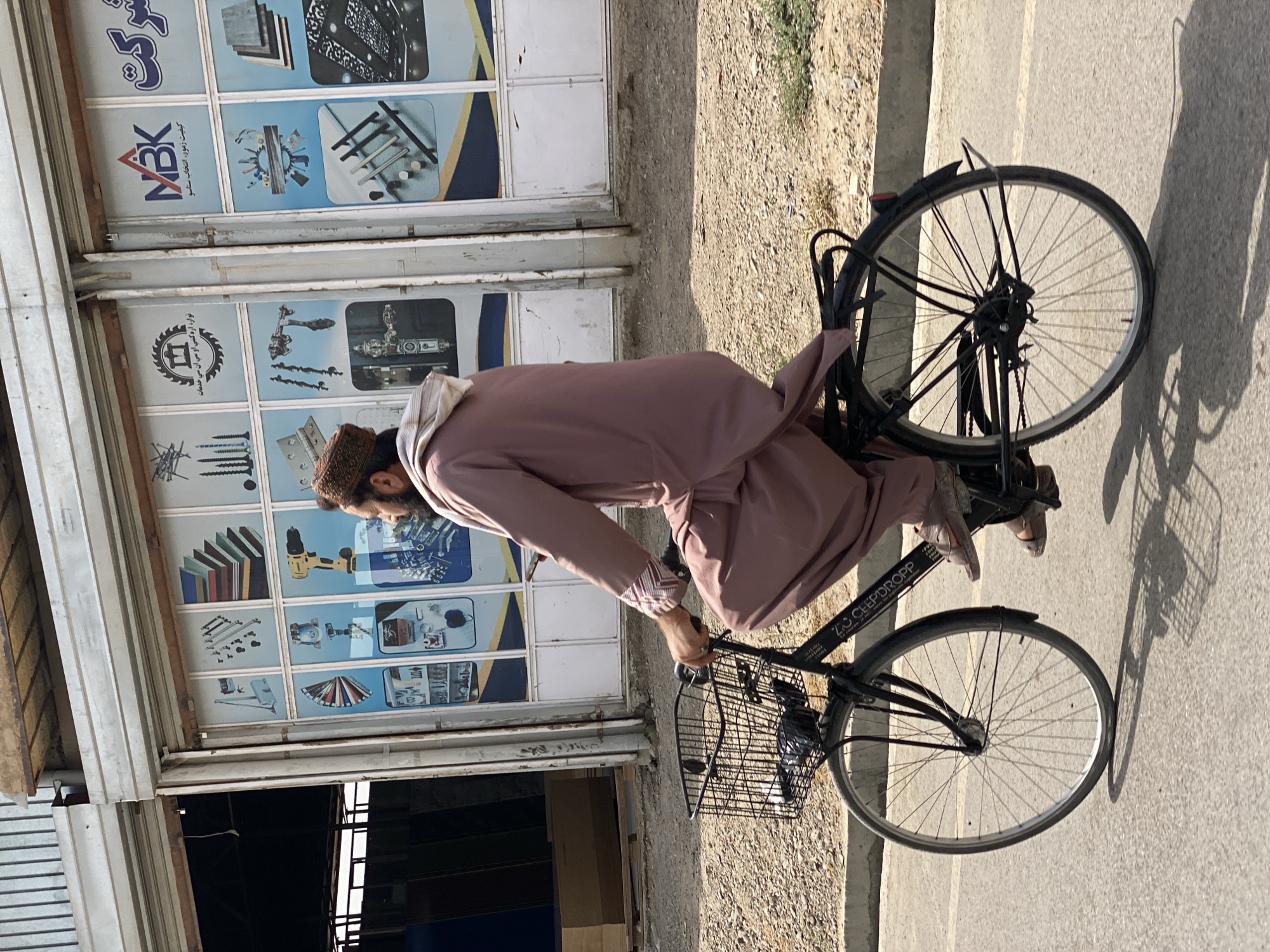 A man wearing a pink tunic and a patterned cap rides a black bicycle with a wire basket on the handlebars. The bicycle has a black frame and spoked wheels. The man is wearing brown sandals and is pedaling the bicycle. In the background, there is a storefront with large windows displaying images of hardware and building materials. The storefront has a metal awning and is located on a dirt road next to a paved street. The environment appears to be a commercial area in Kandahar, Afghanistan. The lighting suggests it is daytime.