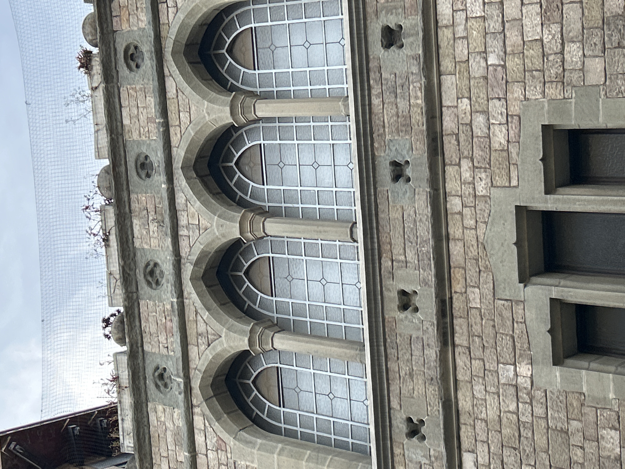 The image displays a detailed section of an ornate stone building facade, seen from an upward angle against an overcast sky. The primary features are a series of four vertically aligned arched windows, each framed by grey stone and separated by short, stout pilasters. These windows are fitted with white frames forming a grid pattern, and some panes feature a distinct diamond motif in their center. The glass appears frosted or opaque, obscuring the interior.
The building's wall is constructed from rough-hewn, light grey and brown stone blocks, laid in an irregular pattern. Embedded within the stone are small, dark, quatrefoil-shaped decorative elements, arranged both horizontally between window sections and vertically in a band to the right. On the far right, a different architectural opening is visible, featuring a more angular, stepped frame in a darker grey stone.
Along the very top edge of the visible building section, several sparse, dry, shrub-like plants with bare branches are growing out of crevices, suggesting the age of the structure. Extending upwards and to the left from this edge is a fine, light-colored mesh netting, likely a protective measure for birds or possibly a construction net. The sky beyond the building is bright but diffused, indicating an overcast daytime setting. No people or specific activities are visible in the static scene. The architectural style suggests an older, possibly historic, structure.