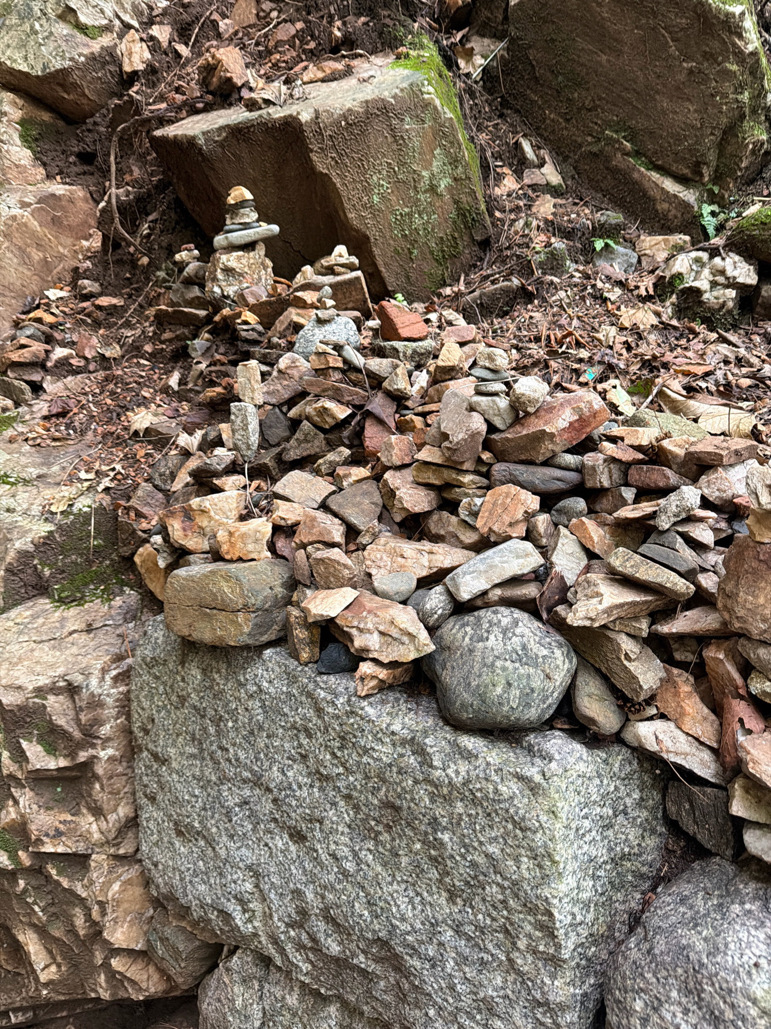 The image shows a natural scene with several rock formations. In the center of the image, there are multiple cairns, which are piles of stones stacked on top of each other. The cairns vary in size and complexity, with some being tall and slender, and others being more spread out. The stones are of various colors and textures, including grey, brown, and reddish hues. The environment appears to be a wooded or rocky area, with fallen leaves and dirt visible on the ground. Large rocks and boulders are present in the background and foreground, some covered with moss. The overall scene is one of a natural landscape with human-made structures (cairns) integrated into it. The location is identified as Dongducheon-si, South Korea.