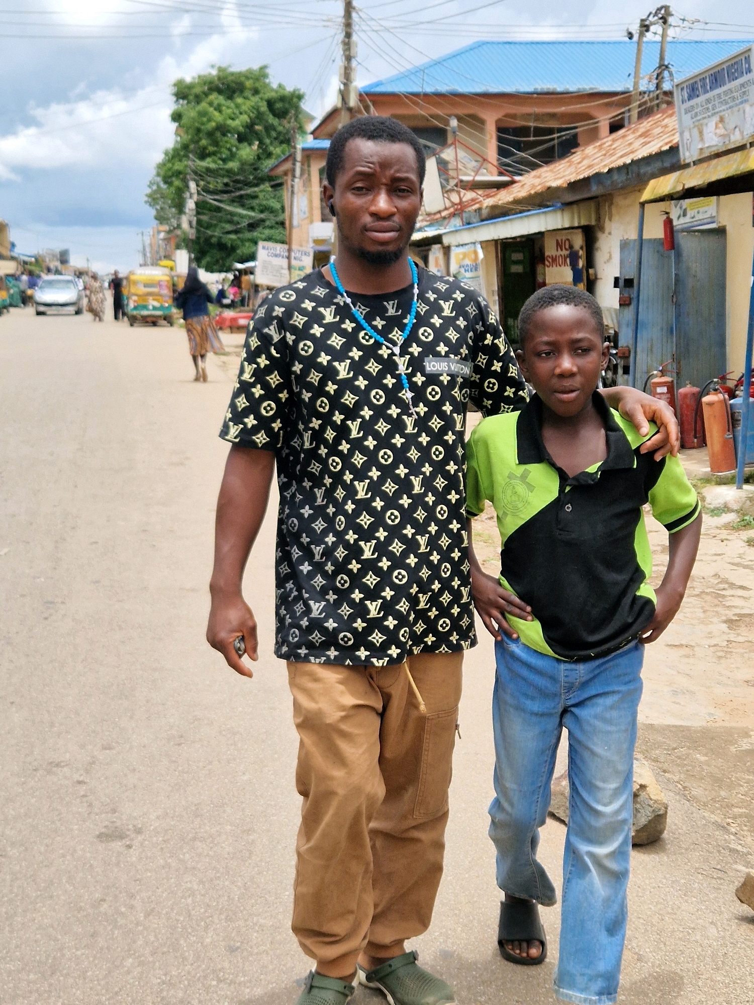 In Jos, Nigeria, a man and a young boy are walking down a paved street. The man, wearing a black t-shirt with a repeating gold pattern and khaki pants, has his left arm around the boy's shoulders. The boy is wearing a green and black polo shirt and blue jeans. They are walking away from the camera. In the background, there are buildings with blue and red roofs, as well as several vehicles, including a car and what appears to be an auto-rickshaw. The sky is overcast with clouds. The scene suggests a typical urban environment in Nigeria, with elements of commerce and everyday life visible.