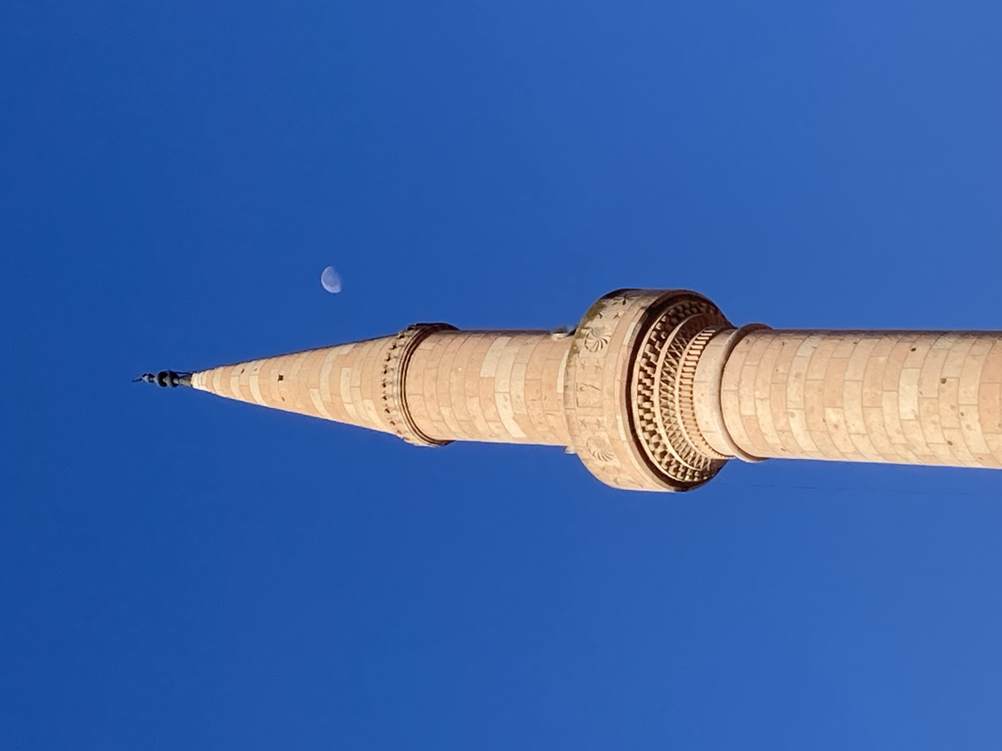 A tall, pale stone minaret rises against a clear, bright blue sky. The minaret is cylindrical at its base and tapers to a pointed, spire-like roof adorned with dark decorative elements. It features intricate architectural details, including decorative bands and carved sections with geometric patterns and what appear to be stylized motifs. A crescent moon, partially illuminated, is visible in the sky to the right of the upper portion of the minaret. The image appears to be a low-angle shot, focusing on the minaret and the expansive sky, with no people or other objects readily apparent in the frame. The scene suggests a clear, sunny day.