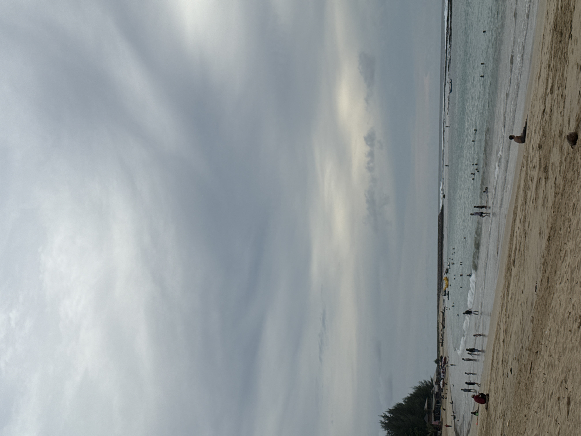 A wide shot shows a beach with a cloudy sky overhead. The sand is a light brown, with footprints and some debris scattered across it. In the foreground, a person is sitting on the sand, facing towards the ocean. Further back, several people are walking along the shoreline and in the shallow water. Some are wading, while others appear to be swimming. A few small waves are breaking on the shore. To the left, a line of trees separates the beach from a cluster of buildings. In the distance, a rocky breakwater extends into the ocean. Several boats, including what appears to be a yellow banana boat, are visible in the water. The overall atmosphere is calm and overcast. This beach is located in the city of Meunasahbale, Indonesia.