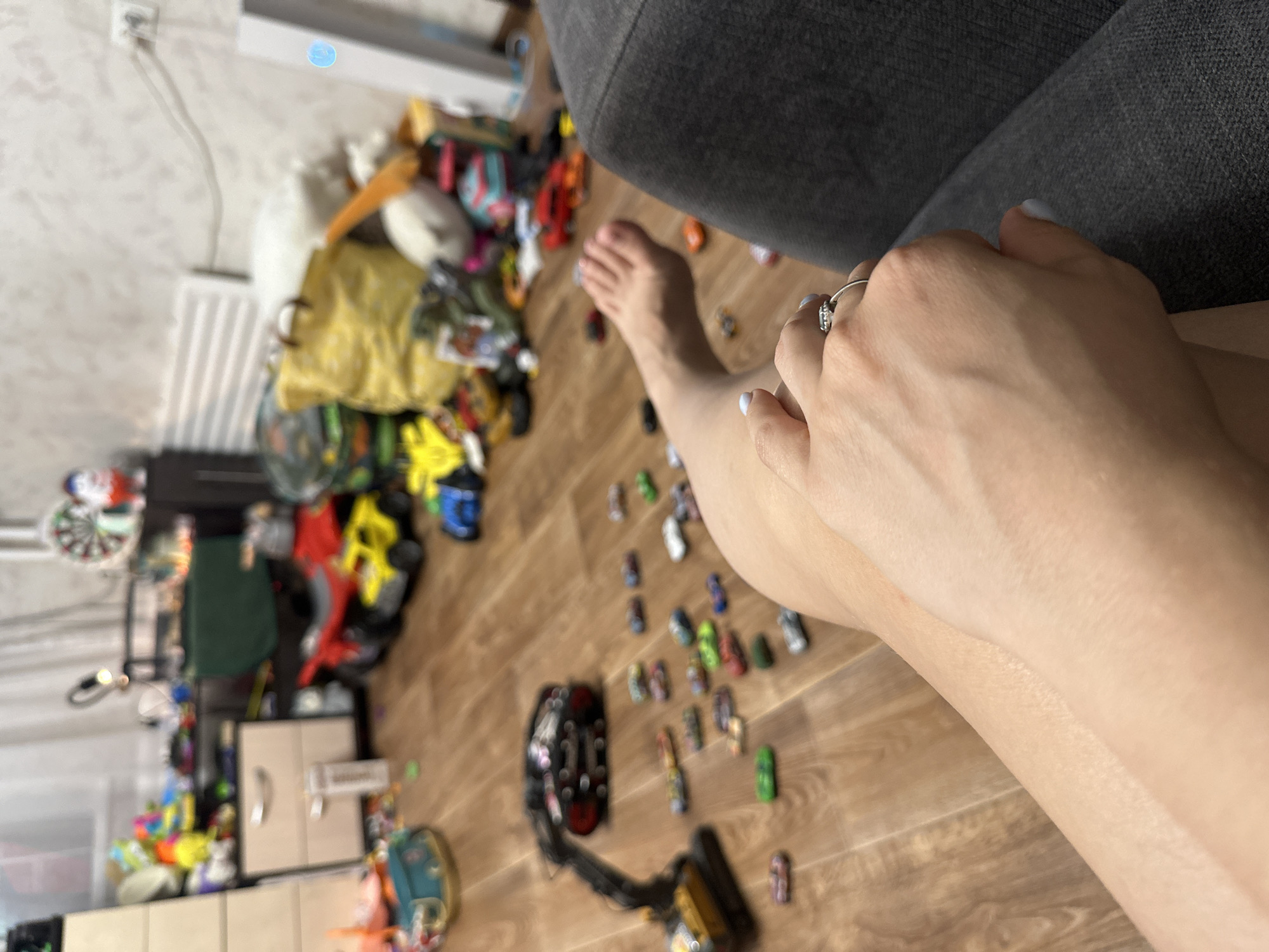 The image depicts a cluttered indoor living space, likely a child's playroom or bedroom, with toys scattered across a wooden floor. In the foreground, a person's legs and hands are visible, suggesting they are seated. The person is wearing a ring on their left hand. The room is filled with various toys, including toy cars, a toy excavator, and larger toys in the background. Furniture such as a chest of drawers and shelves are also present, contributing to the overall disarray. The lighting suggests it is daytime, with artificial light also contributing. The location is described as Novoaltaysk, Russia.