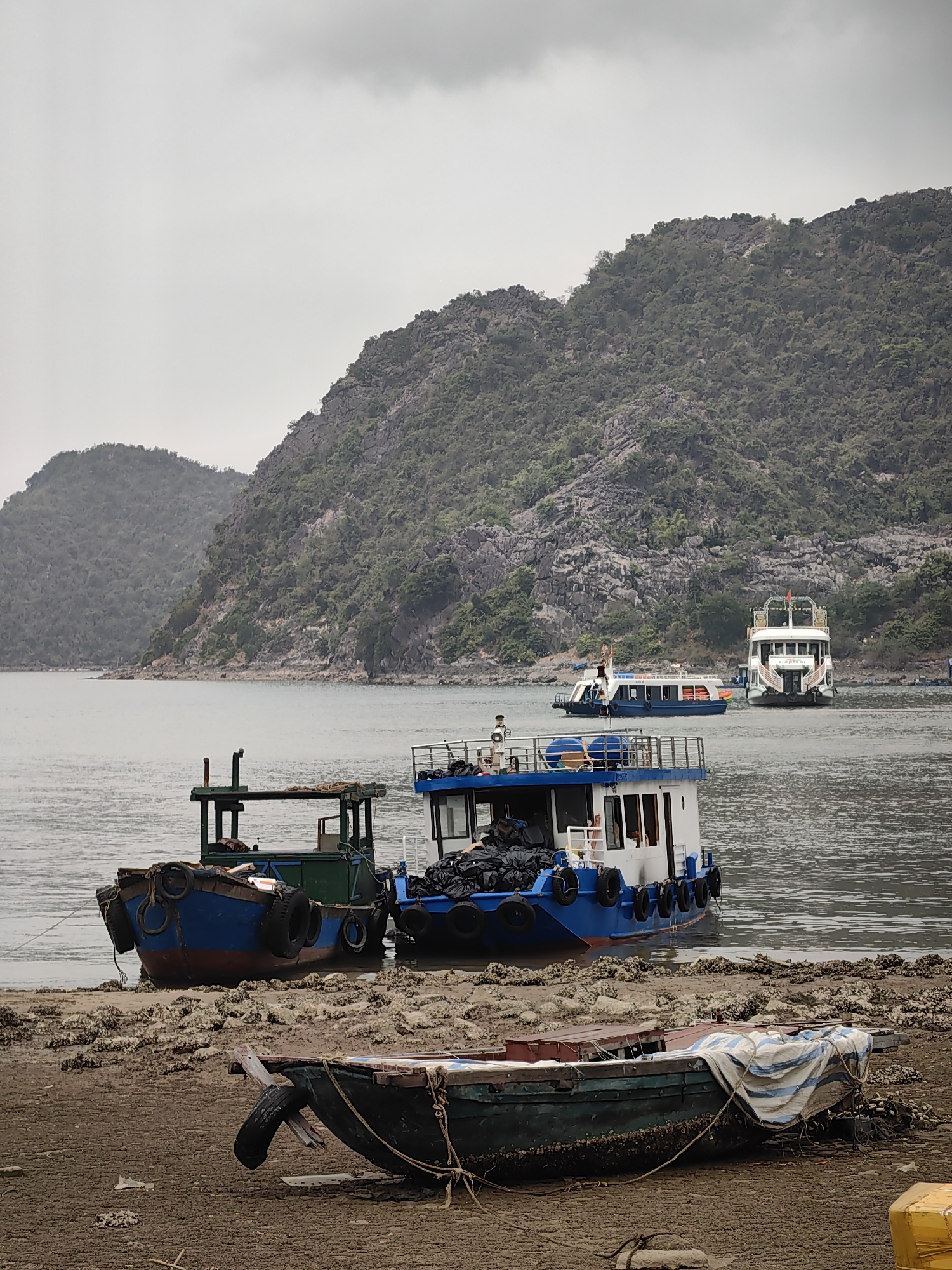 The image shows several boats near the shore of a body of water. In the foreground, a small, weathered boat rests on the beach, partially covered by a striped tarp. Behind it, two larger blue and green boats are partially beached with tires used as bumpers. In the background, there are two more large white boats, indicating possible tourist activity. The background also includes green, rocky hills or mountains, and the sky is overcast, suggesting a cloudy day. The location is likely in the city of Hải Phòng, Vietnam, given the landscape and type of boats. The scene depicts a harbor or bay area where boats are docked or resting.