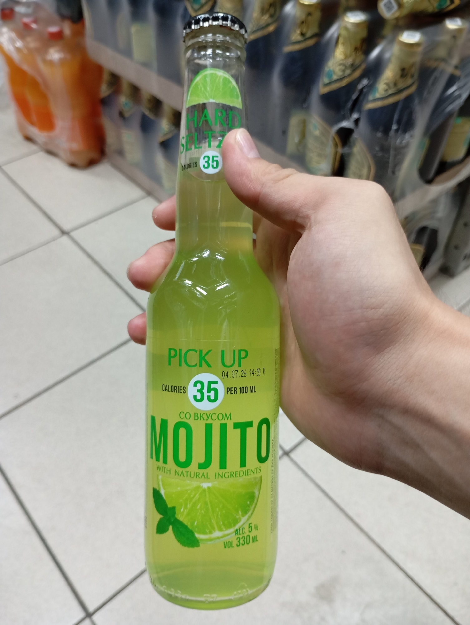 A person holds a glass bottle of "PICK UP MOJITO" drink in a store. The bottle is mostly filled with a light green liquid and has a label with text and graphics. The label indicates "CALORIES 35 PER 100 ML" and features a lime slice and mint leaves. The background shows shelves with other beverages. The scene appears to be indoors, possibly a supermarket or convenience store, given the tiled floor and rows of products. The person's hand is visible holding the bottle. The lighting suggests it is daytime.