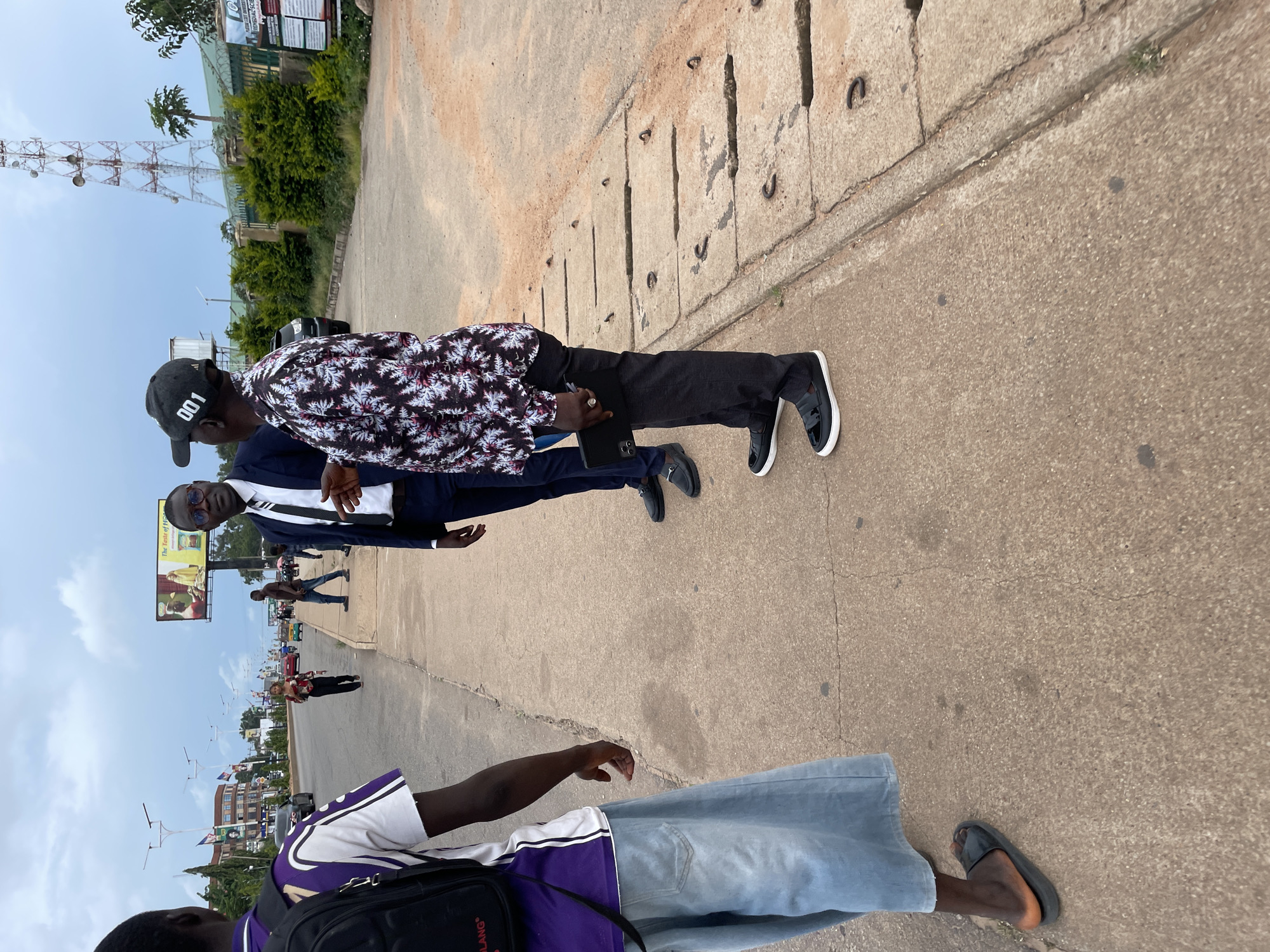 The image captures a street scene in Jos, Nigeria. In the foreground, three individuals are visible. The person on the far left is facing away from the camera, wearing a purple and white shirt and light blue wide-leg pants, with a black backpack. To their right, two men are engaged in conversation. The man in the middle is wearing a dark suit, white shirt, and tie, with glasses. The man on the right is wearing a patterned shirt, dark pants, and a black baseball cap with "001" printed on it. He is holding a dark-colored electronic device, possibly a tablet or large phone, in his left hand. The environment appears to be a sidewalk next to a road. In the background, there are vehicles, buildings, streetlights, flags, and a prominent telecommunications tower. A billboard advertising "The Taste of H..." is also visible. The sky is overcast, suggesting cloudy weather. The ground is paved with concrete and a reddish-brown dirt substance. There are visible drainage covers along the edge of the sidewalk.