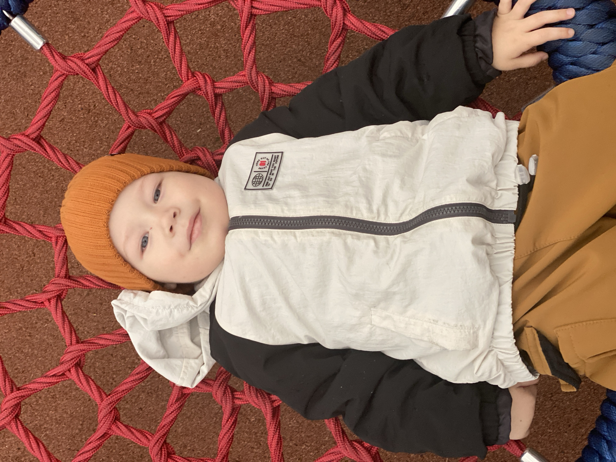 A young child, appearing to be a toddler, is lying comfortably on a large red rope net, likely part of a playground swing or hammock. The child is positioned centrally, looking directly at the camera with a gentle, content expression and a slight smile. They have fair skin, light hair visible under a hat, and bright blue eyes.
The child is dressed in an orange knitted beanie, a two-tone black and white long-sleeved jacket, and mustard or khaki-colored pants. The jacket features a prominent rectangular patch on the left chest. Text on this patch reads "THINK OUT OF" (partially visible), displays a globe icon, and includes "FILA" below the globe. Vertically arranged text on the right side of the patch states, "FIND OUT WHAT YOU WANT TO DO IN THE FUTURE". A dark grey zipper is visible across the front of the jacket.
The red rope net, intricately woven, forms the surface the child is resting on. Beneath the net is a reddish-brown granular ground, typical of playground surfacing. Thick blue ropes are visible in the background, securing the net, with a silver metal connector visible where the red net meets the blue rope. The setting is an outdoor playground during daytime in Sochi, Russia, suggesting mild to cool weather given the child's attire.